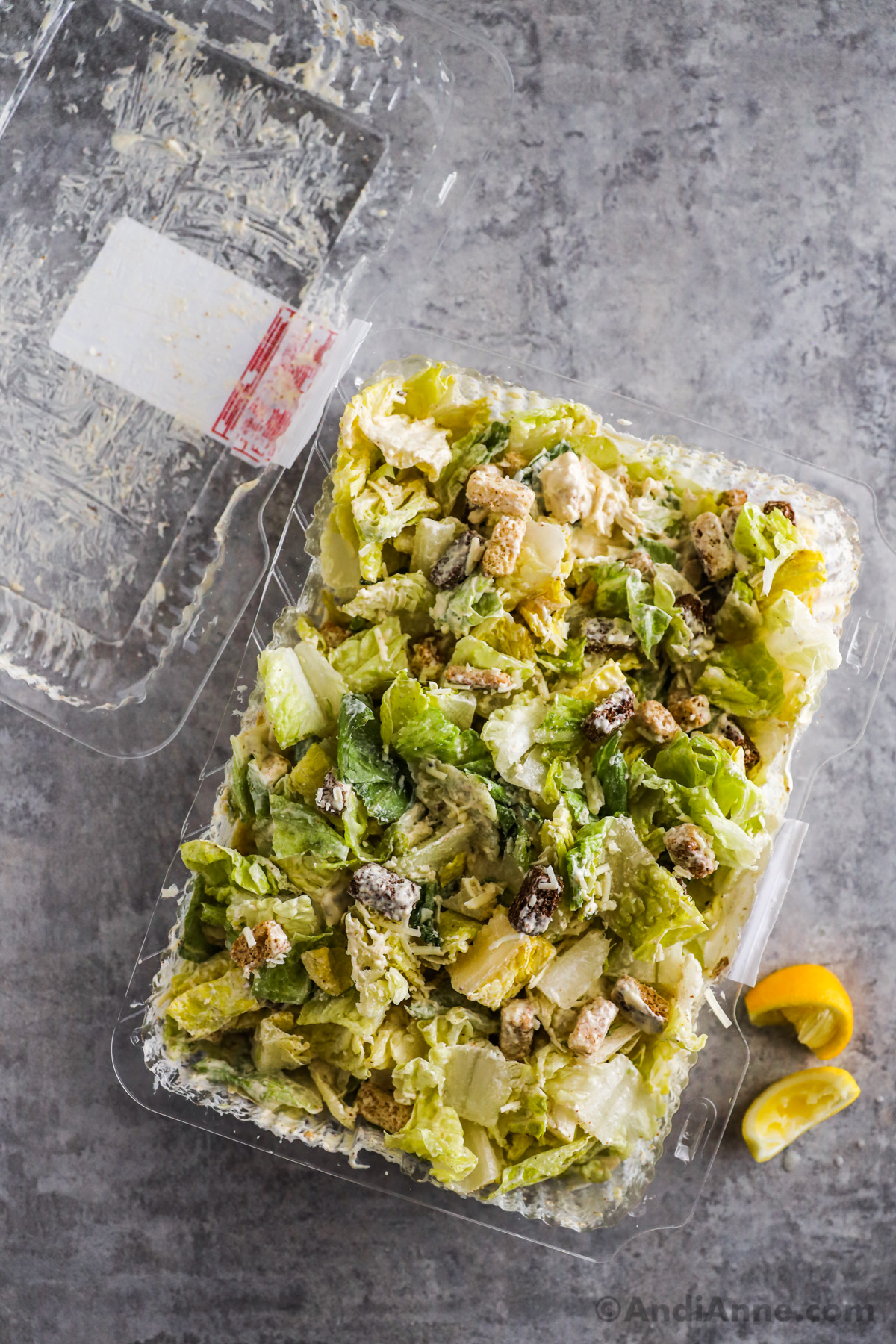 A Costco caesar salad in a plastic container with lemon wedges and a lid beside it.