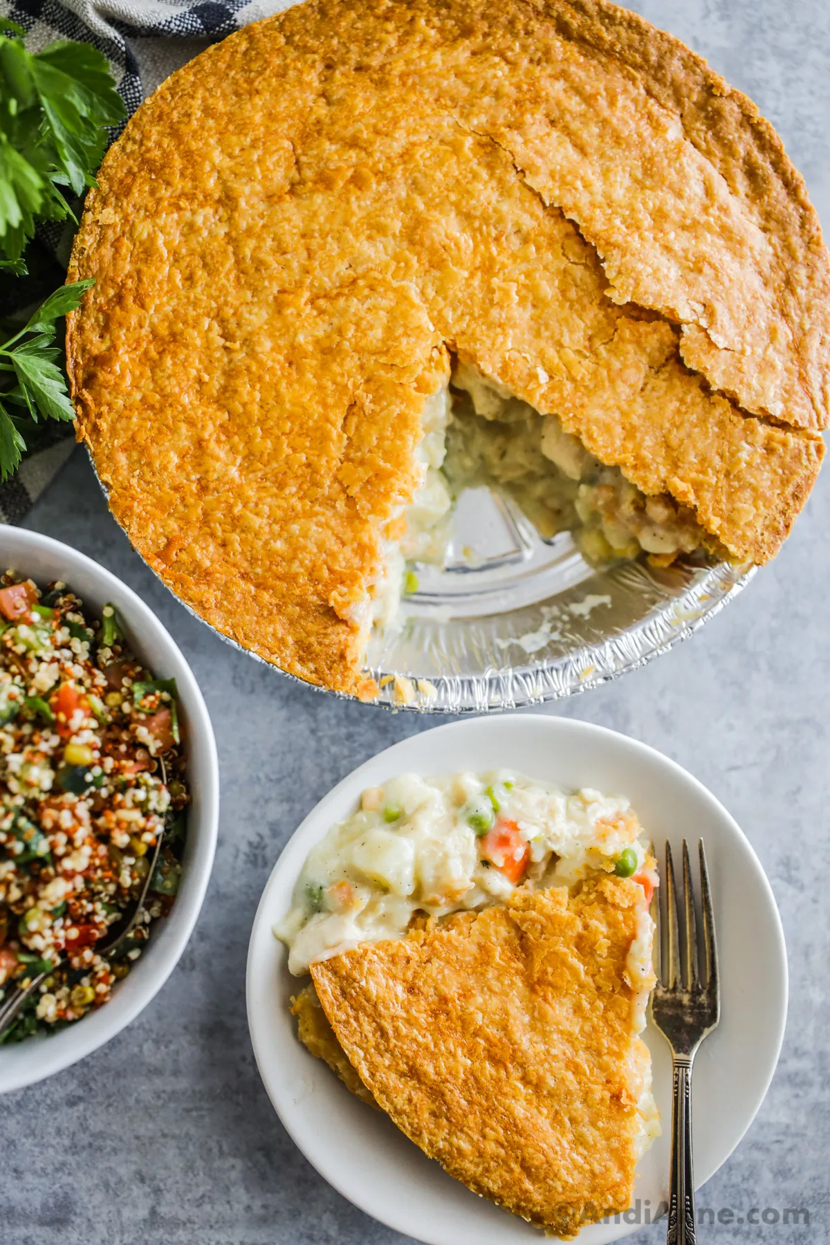 A slice of costco chicken pot pie on a plate with the full pie and a bowl of quinoa salad beside it.