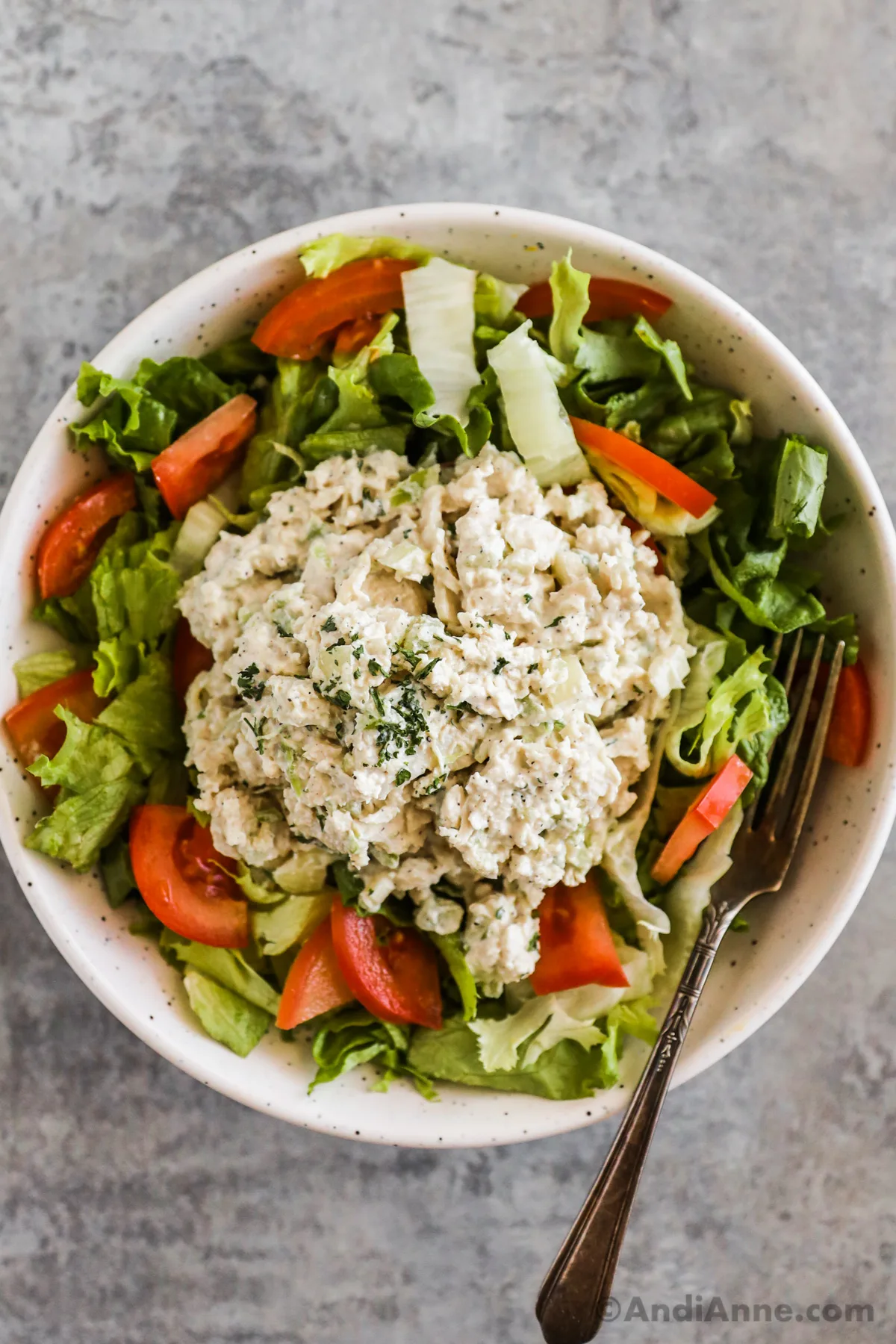 Chicken salad overtop of greens and tomatoes in a white bowl with a fork.