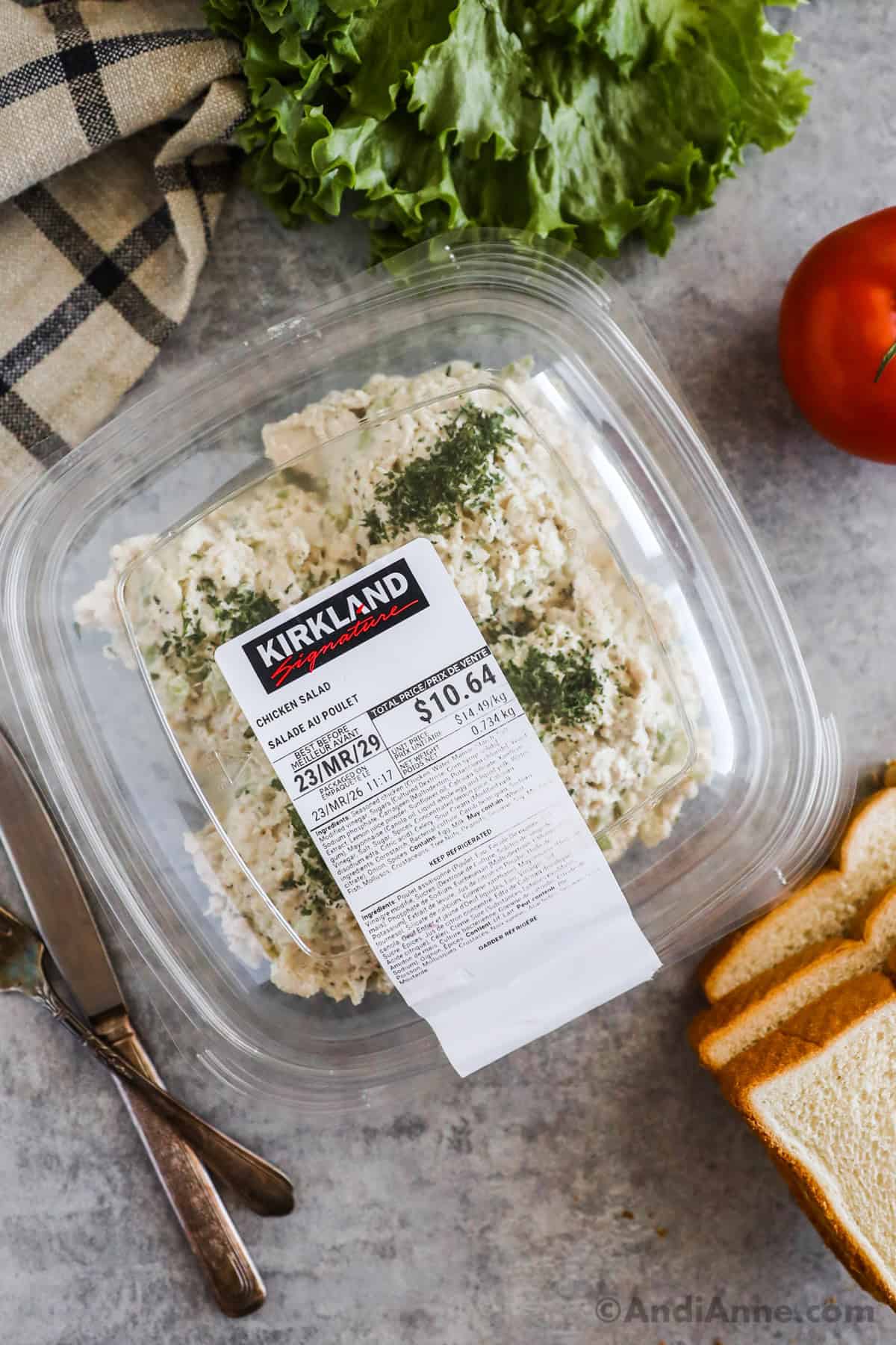 A plastic container of costco kirkland signature chicken salad with lettuce, bread slices and a tomato surrounding it.