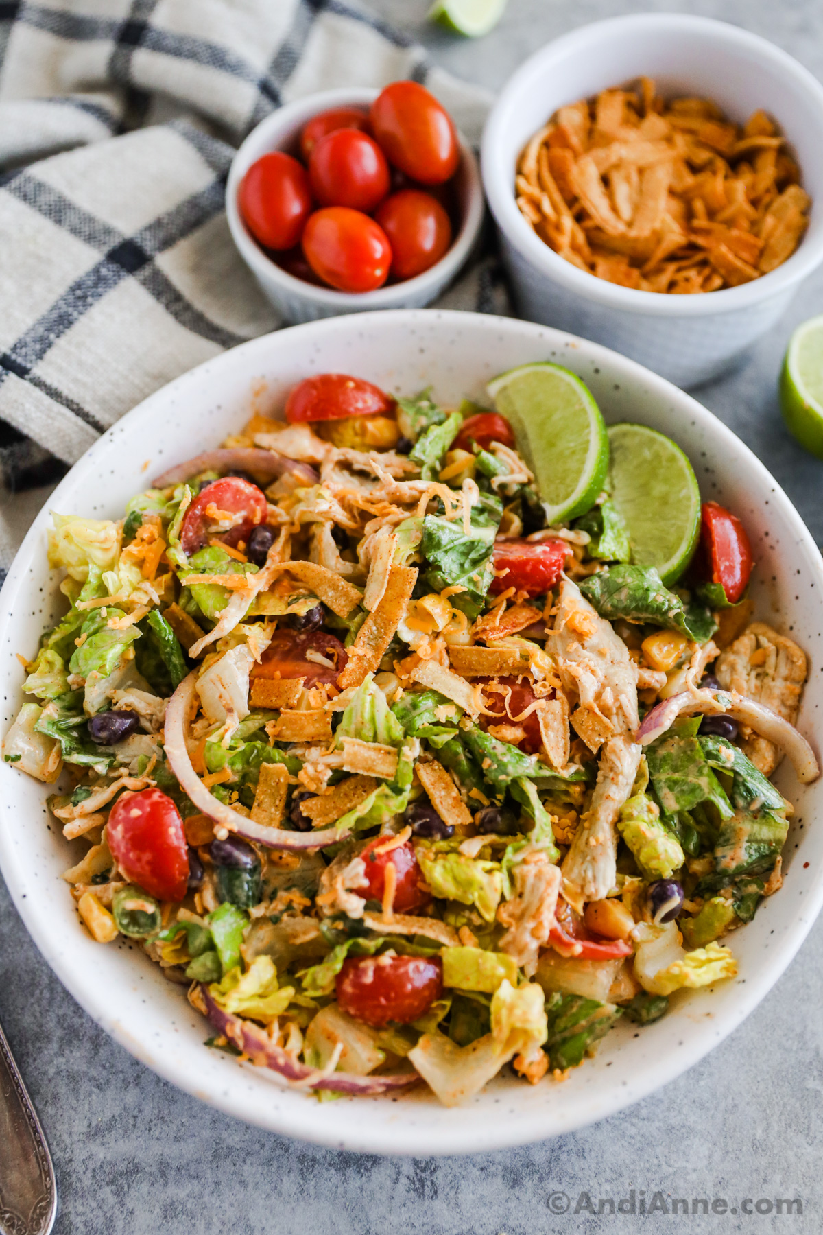 Chicken taco salad in a white bowl with small bowls of cherry tomatoes and tortilla strips beside it.
