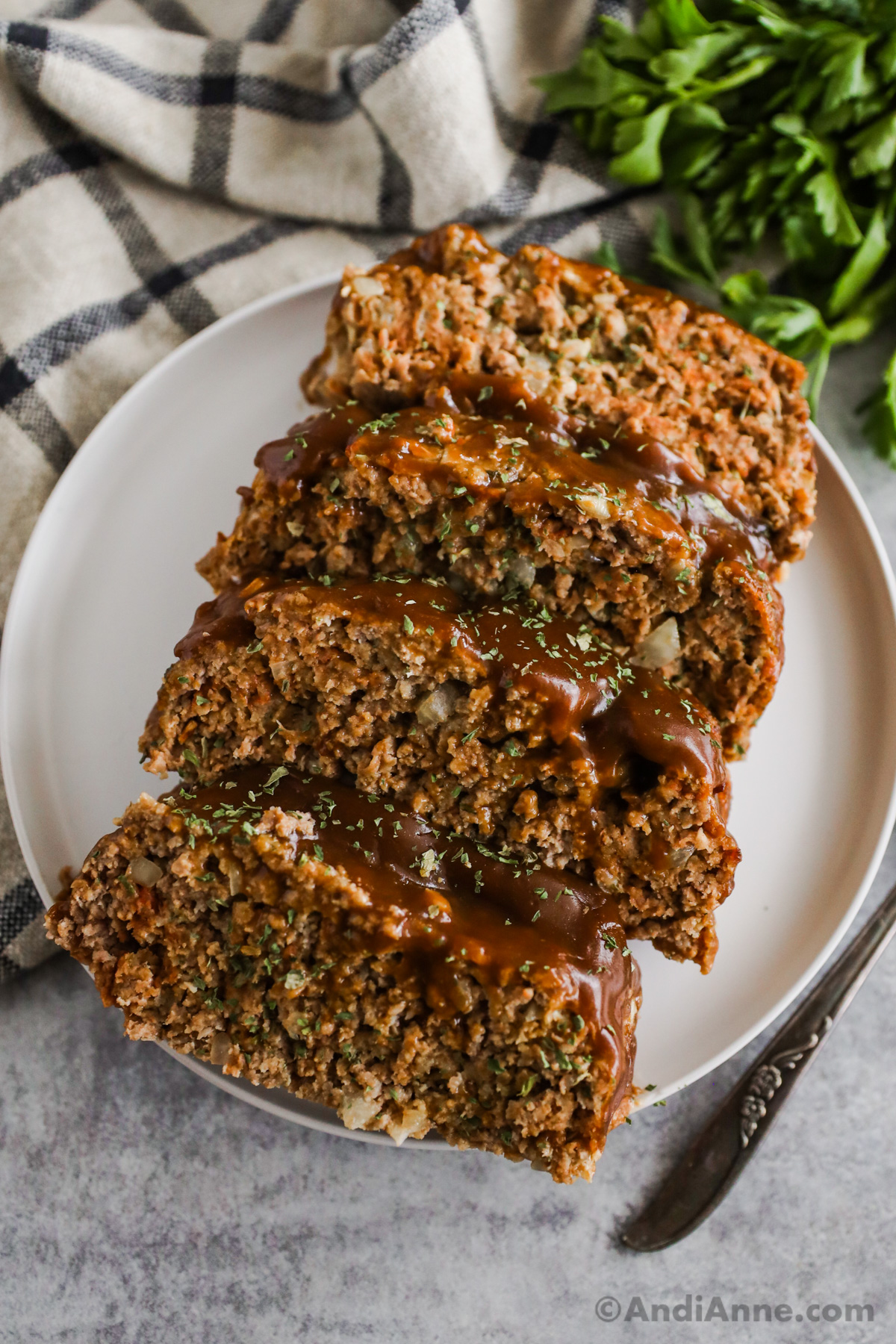 Slices of meatloaf with brown gravy on a white plate with a kitchen towel and parsley in the background.