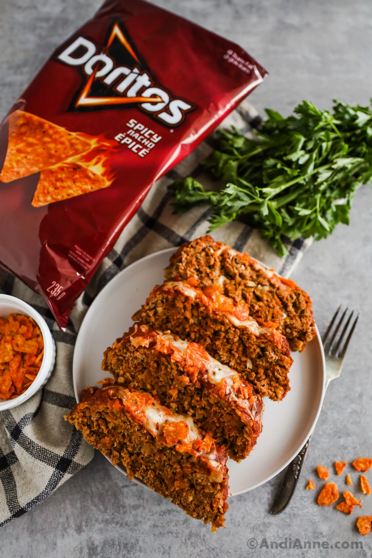 Slices of taco meatloaf on a white plate with a bag of Dorito nacho chips, parsley, fork and kitchen towel in background.