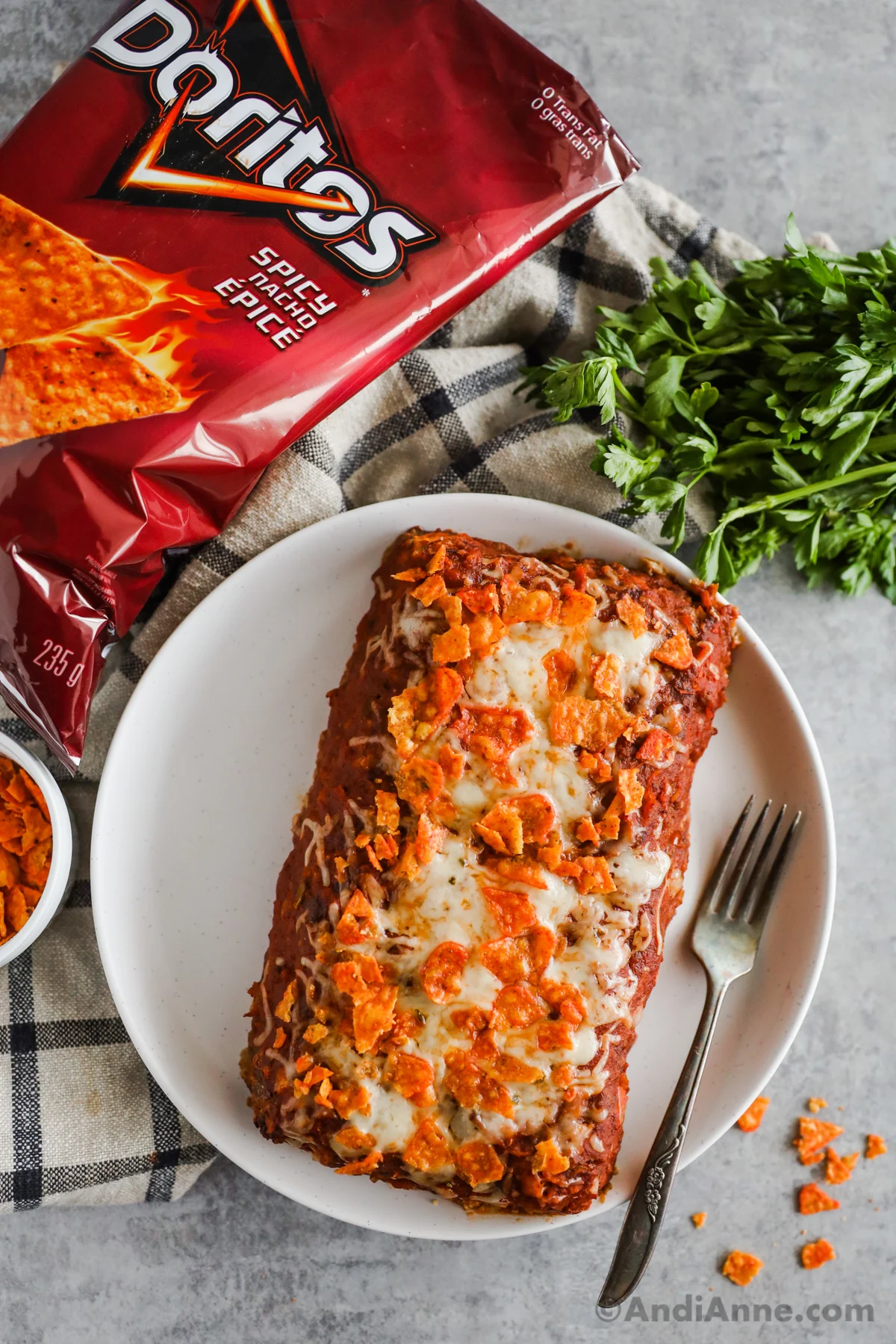 Taco meatloaf on a white plate with a fork. A bag of Doritos, parsley and kitchen towel surround it.