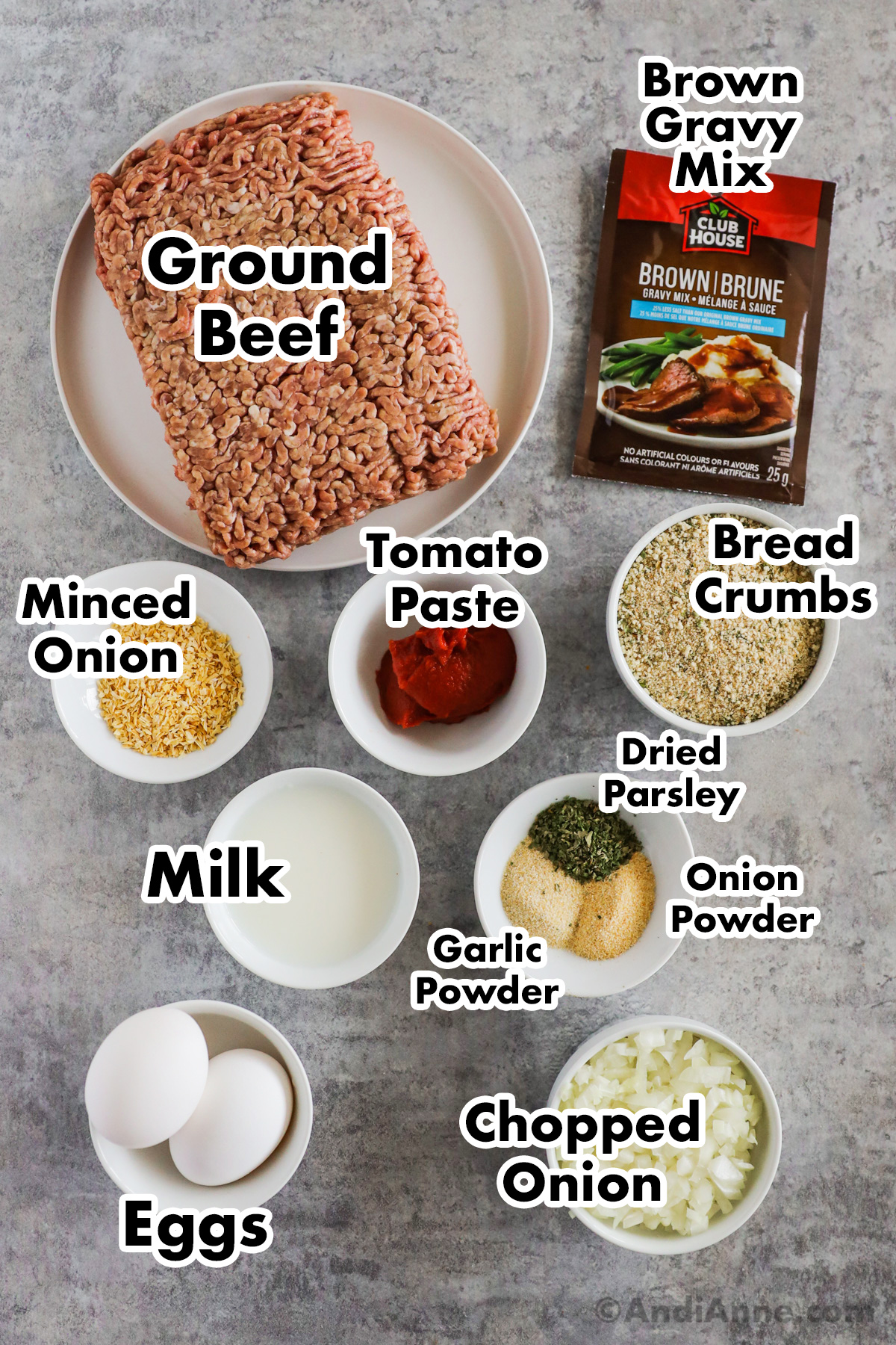 Recipe ingredients on a counter including raw ground beef, packet of brown gravy, bowls of minced onion, tomato paste, bread crumbs, milk, spices, eggs and chopped onion.