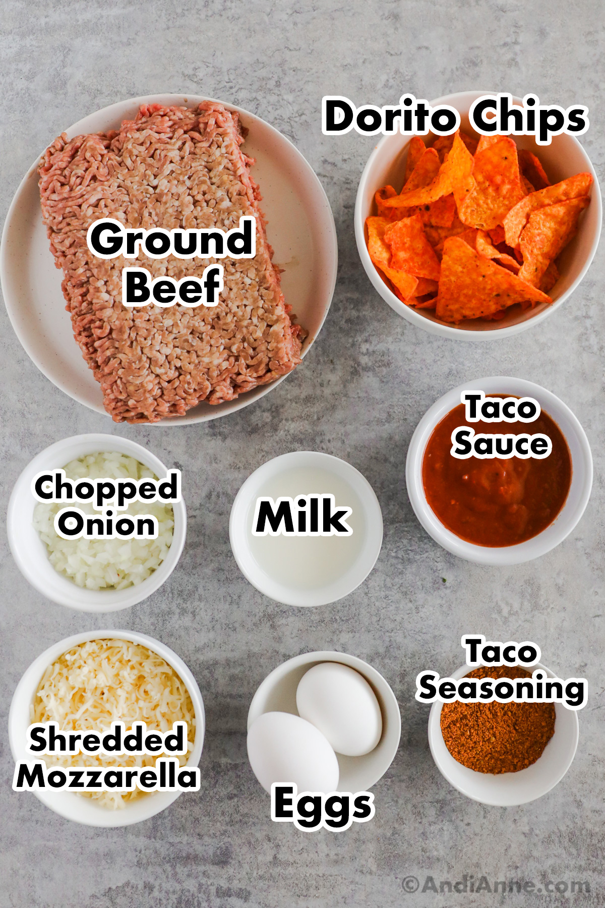 Recipe ingredients on the counter including raw lean ground beef, bowl of Dorito chips, bowls of chopped onion, milk, taco sauce, shredded mozzarella, eggs and taco seasoning.