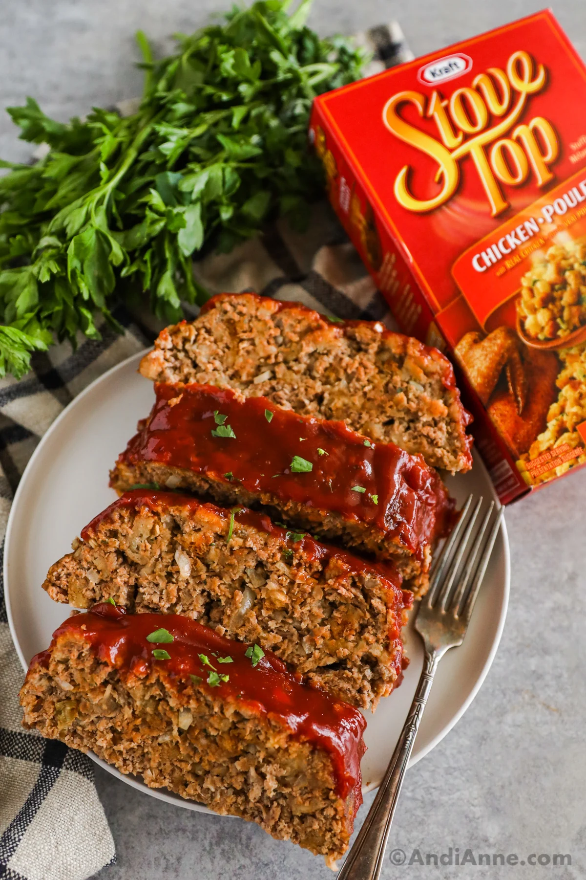 Slices of meatloaf on a plate with a fork and box of stove top stuffing and bunch of parsley beside it.