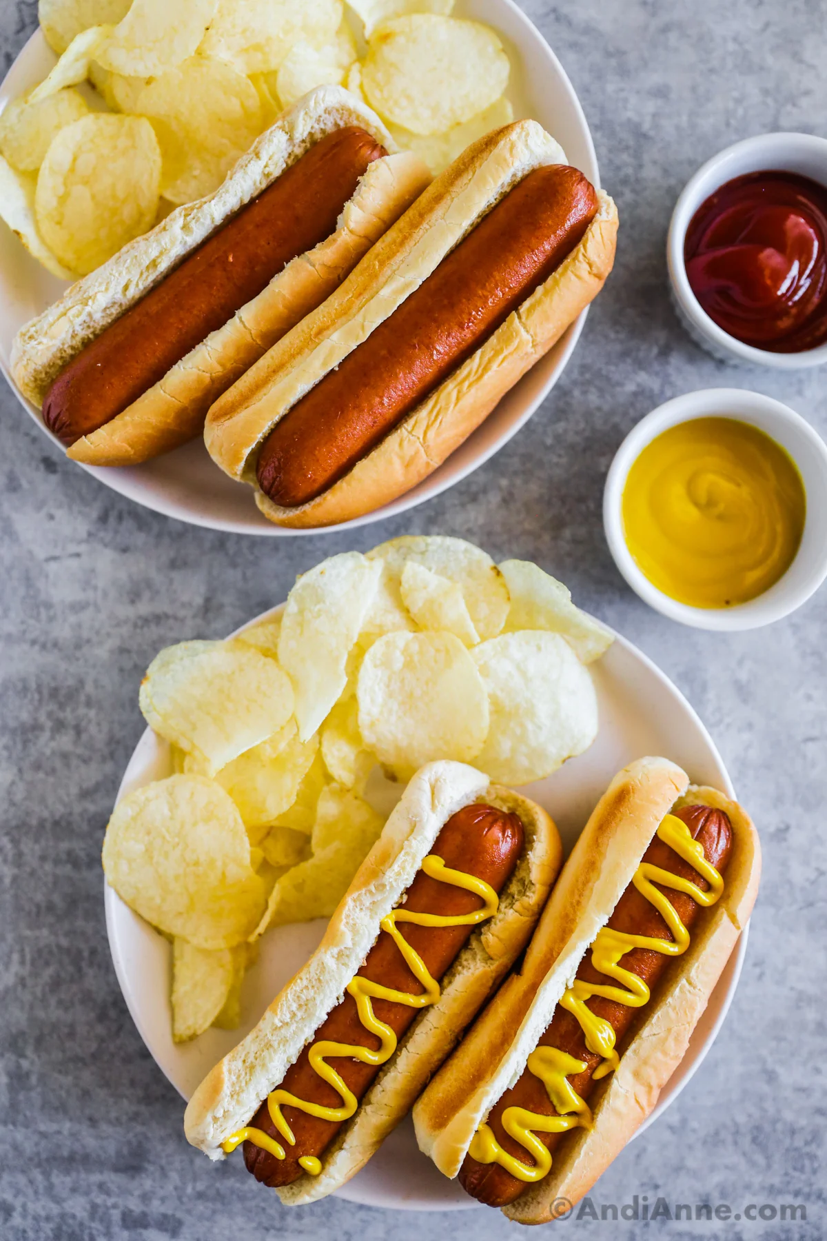 Two plates of with hot dogs and buns and chips, one with mustard on top. Two small bowls of ketchup and mustard beside.