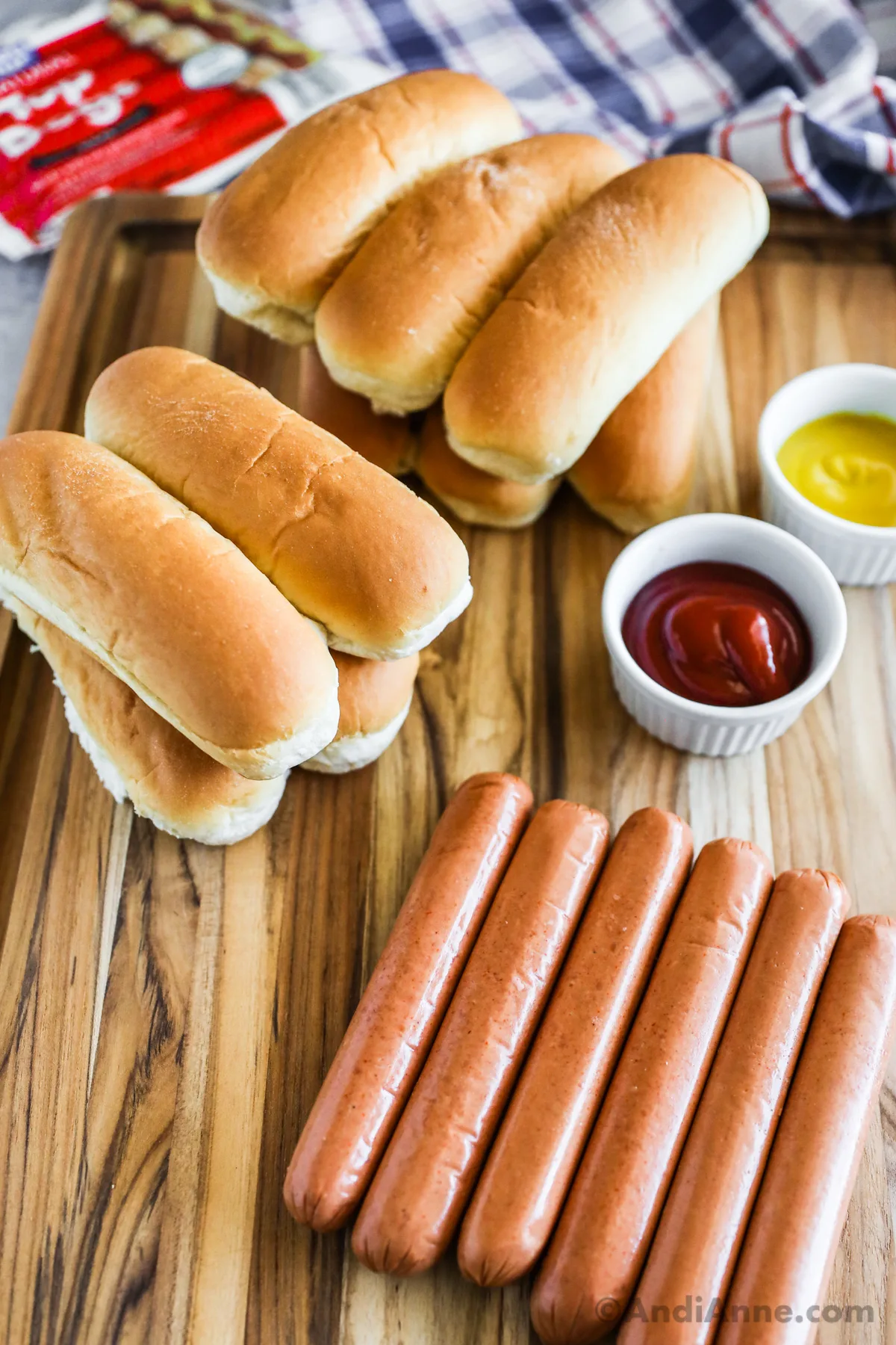 A stack of buns, hot dogs and small bowls of ketchup and mustard.