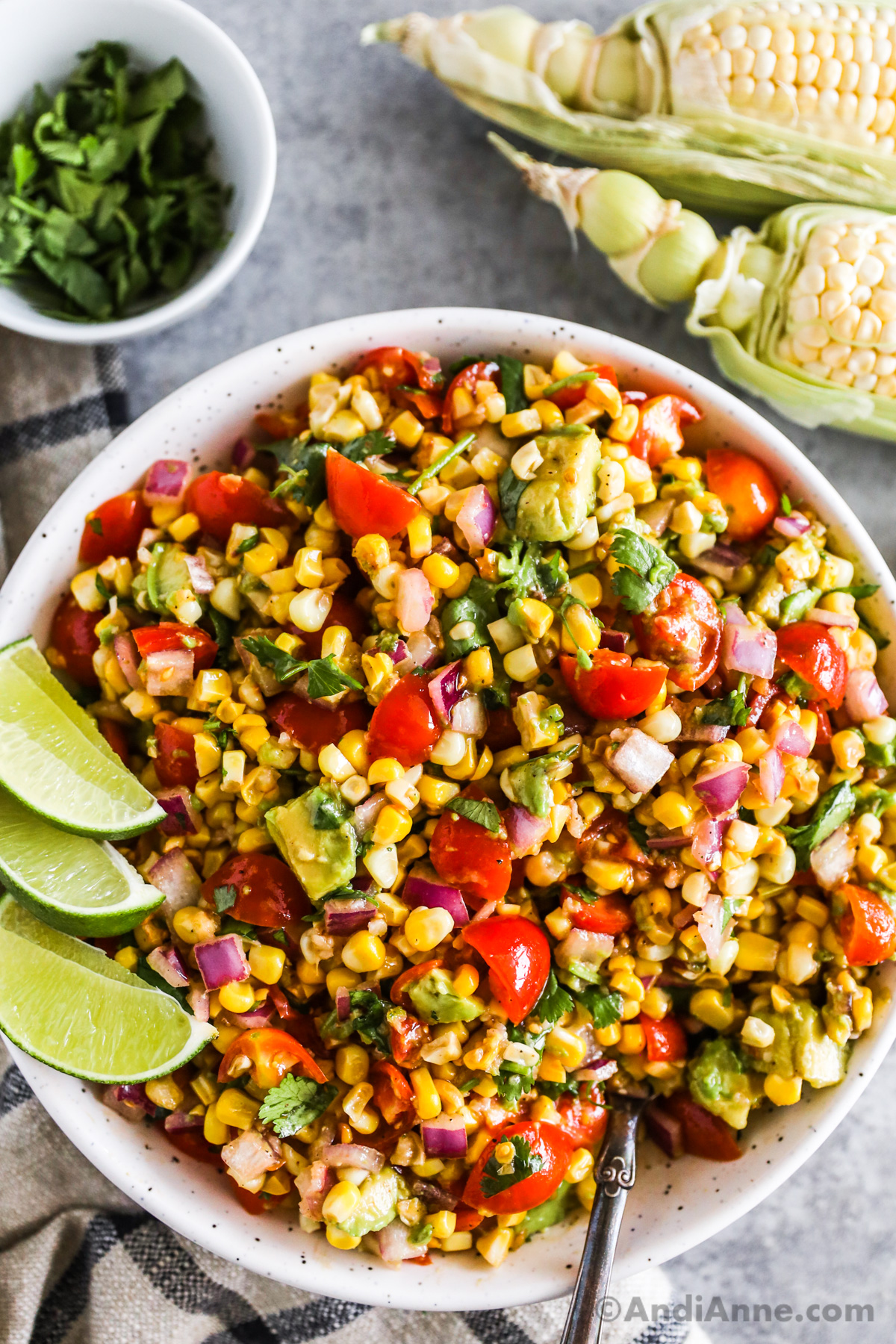 A bowl of corn tomato avocado salad with three lime wedges.