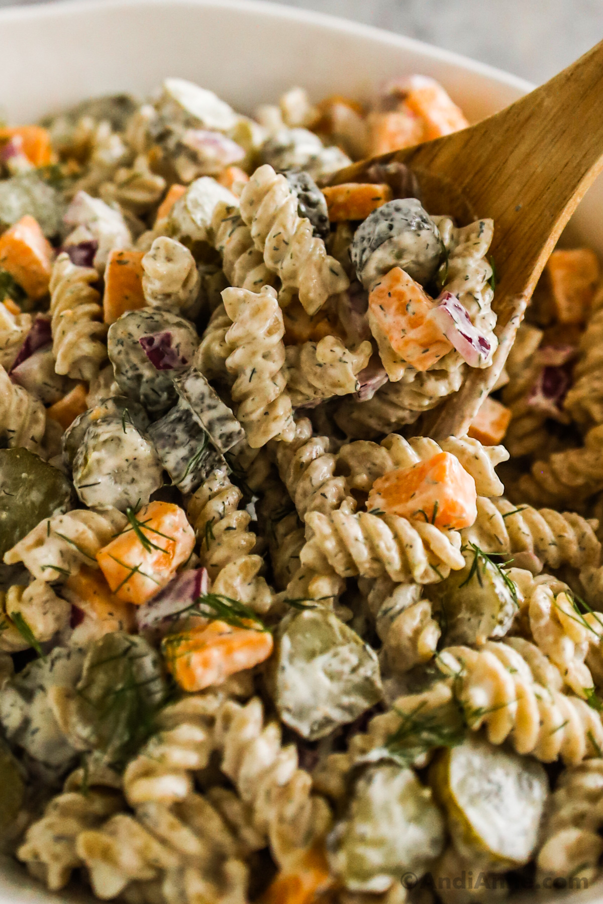 Close up of rotini pasta, cheese, dill pickles and onion, all in a creamy dressing of the pasta salad.