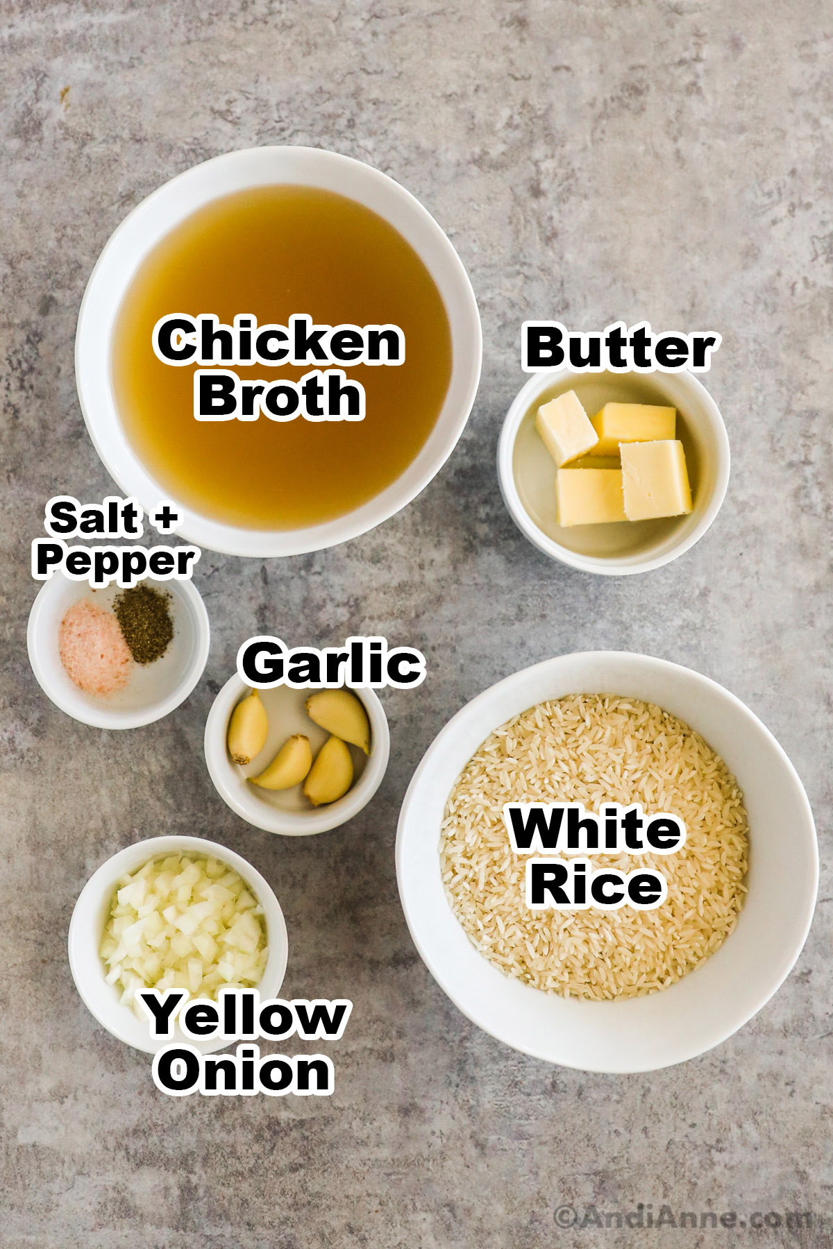 Recipe ingredients on the counter including bowl of chicken broth, bowl of cubed butter, bowls of garlic cloves, uncooked white rice, chopped yellow onion.