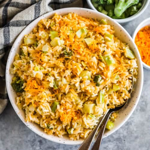 Broccoli cheddar rice in a white bowl with small bowls of broccoli and kitchen towel beside.