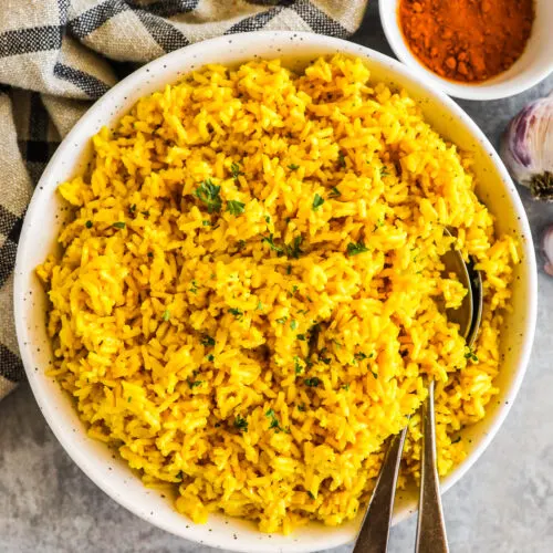 A white bowl of turmeric rice with spoons and a small bowl of turmeric powder.