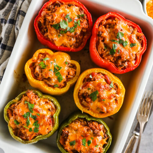 A white baking dish with six baked stuffed peppers topped with melted cheese.