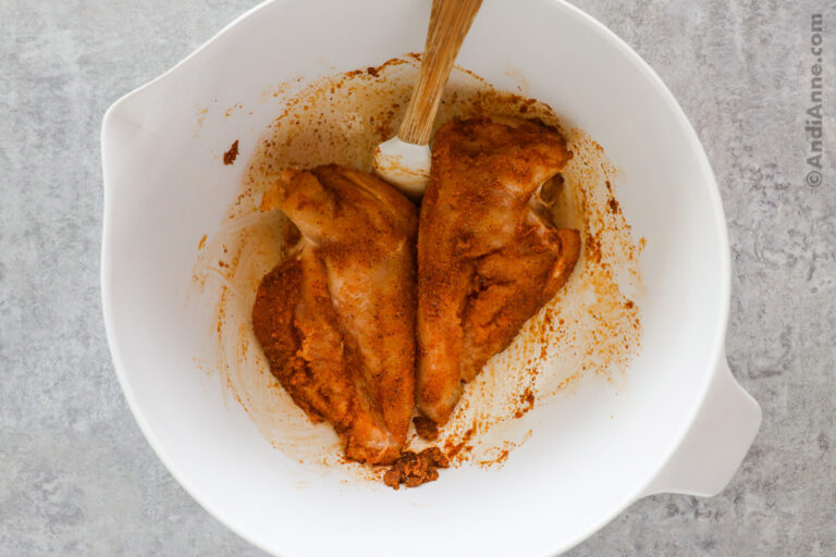 Two raw chicken breasts tossed in spices in a white bowl with a spatula.
