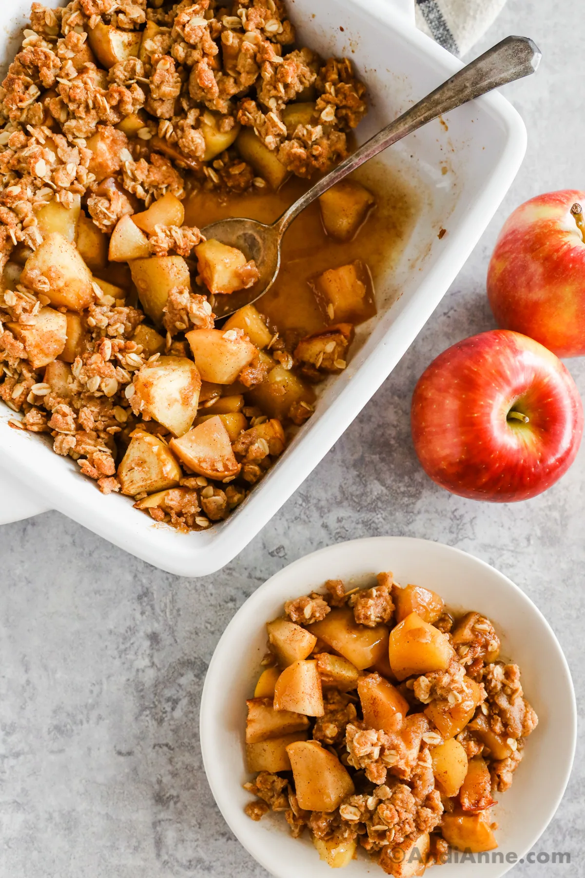A white baking dish with cinnamon apple crisp recipe and a spoon. A serving of the apple crisp on a plate is beside the dish.