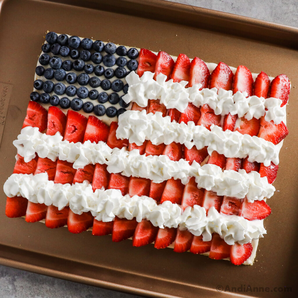 American flag design fruit pizza made from sugar cookie crust, cream cheese frosting, sliced strawberries, whipped cream and blueberries.
