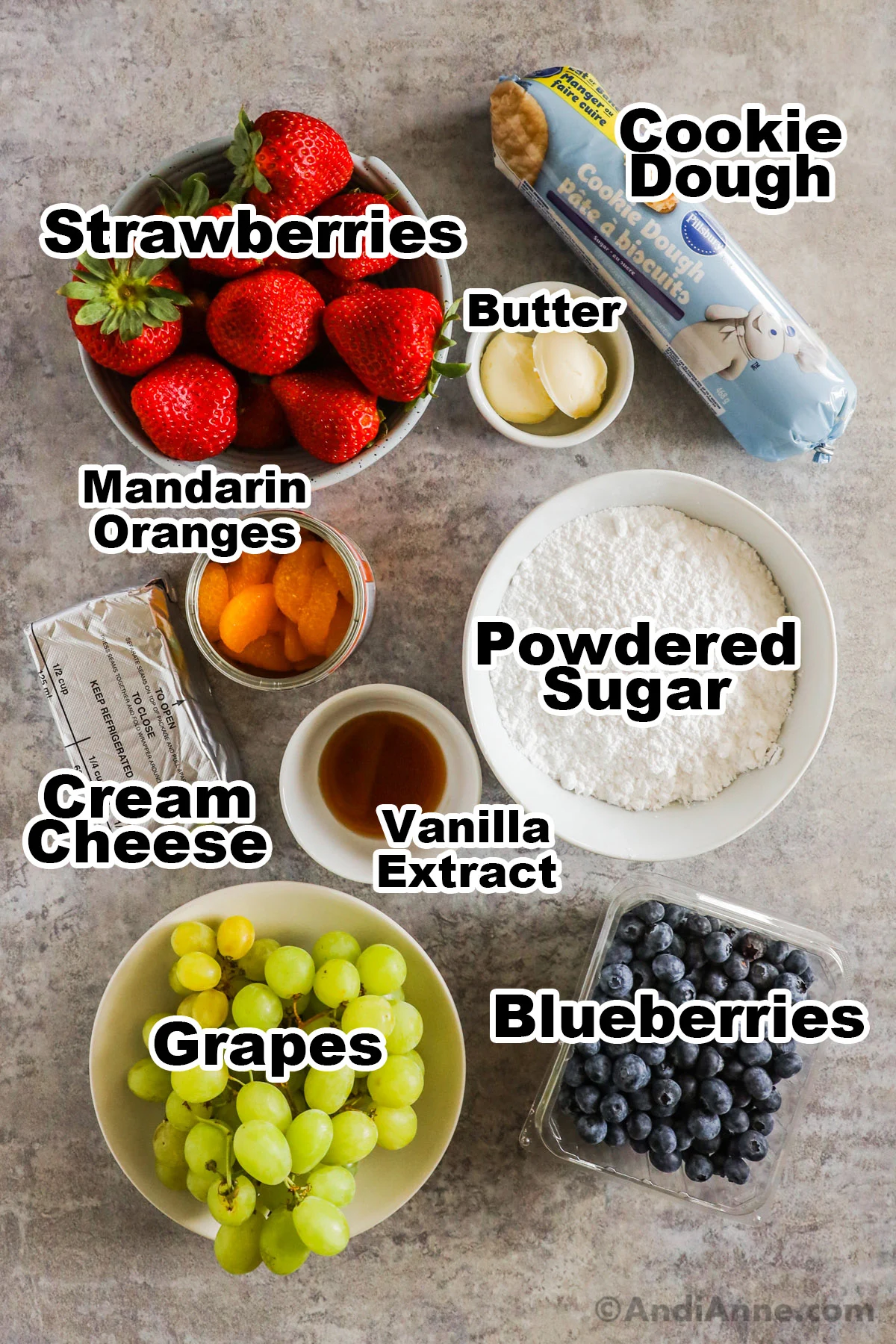 Recipe ingredients on the counter including bowls of fresh strawberries, grapes, blueberries, some cream cheese, refrigerated cookie dough, powdered sugar and vanilla extract.