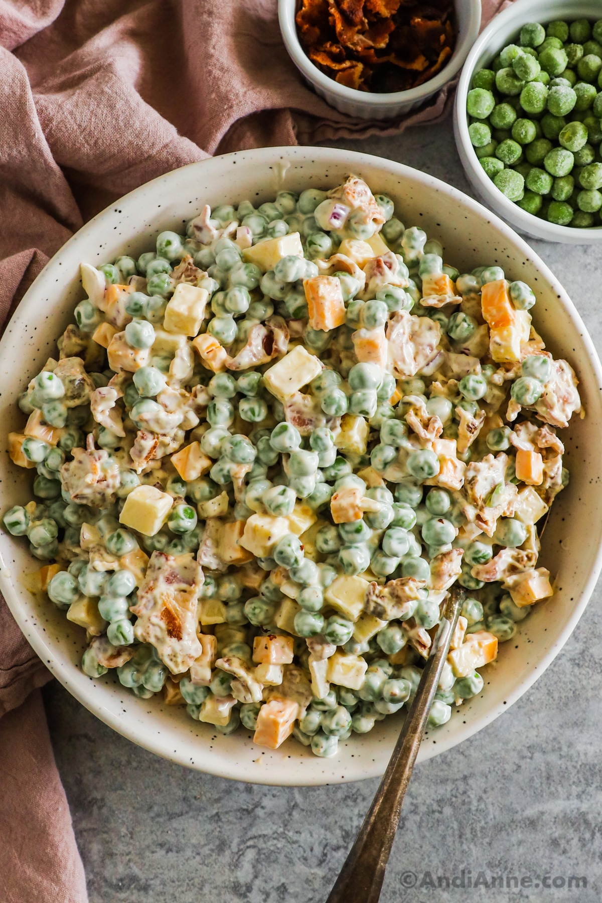 Creamy pea salad recipe with cubed cheese, bacon and onion in a bowl. Small bowls of crumbled bacon and frozen peas surround the bowl.