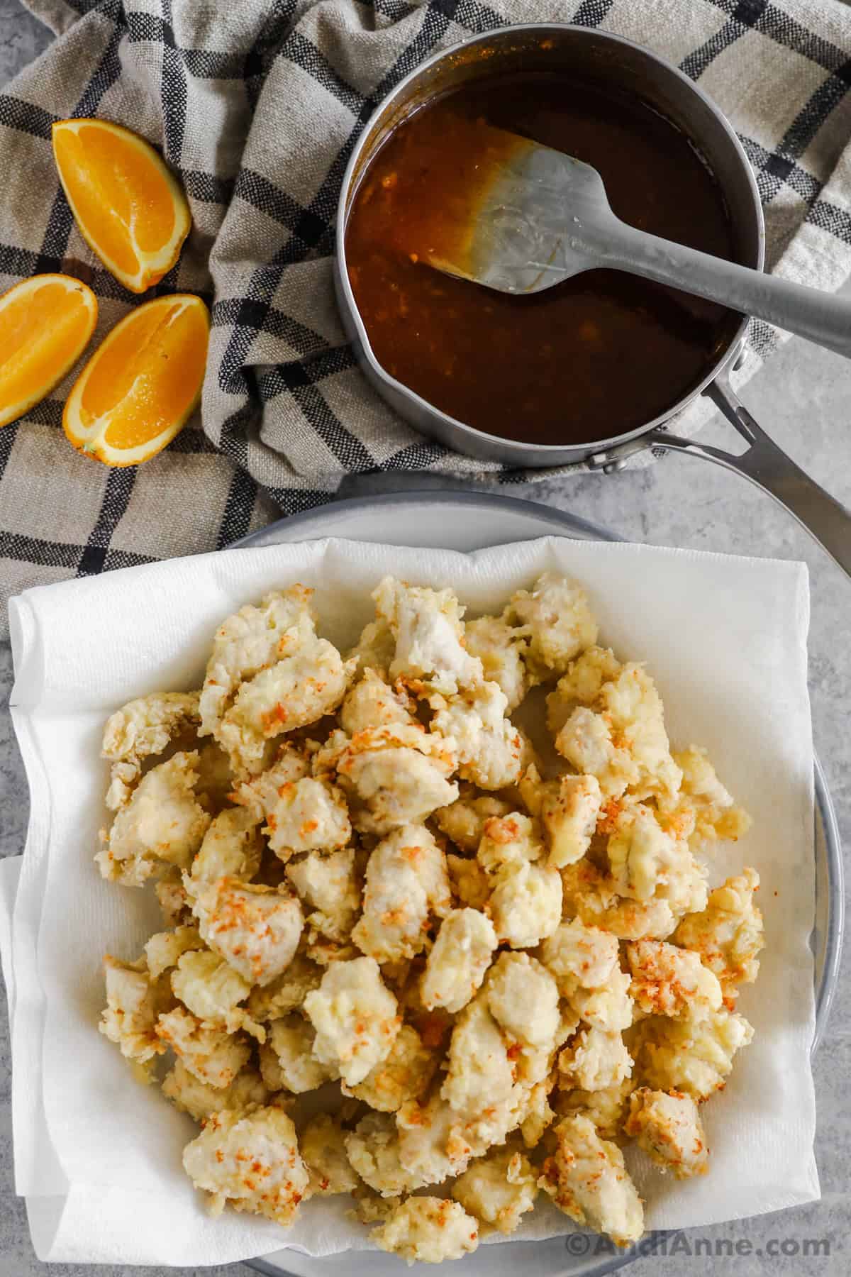 Cooked chicken pieces on a plate with a saucepan with orange sauce and orange slices beside it.
