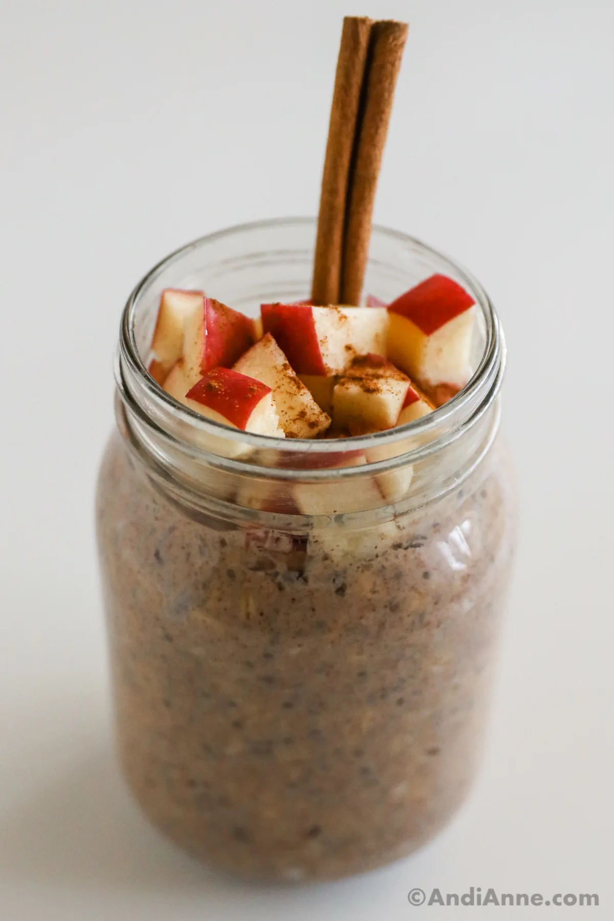 A mason jar with apple cinnamon overnight oats topped with apple chunks and a cinnamon stick.