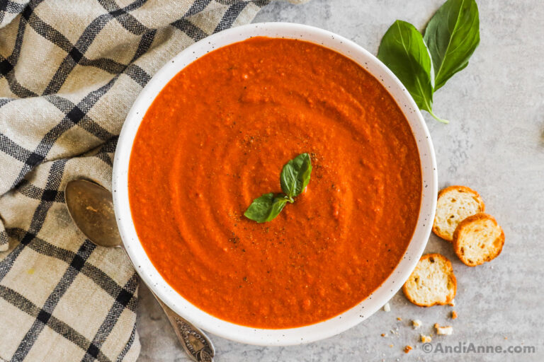 Creamy tomato soup with a basil leaf garnish in a white bowl with crackers and more fresh basil beside.