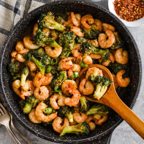 Shrimp and broccoli in honey garlic sauce in a frying pan with a wood spoon. Bowls of green onion and red pepper flakes beside pan.