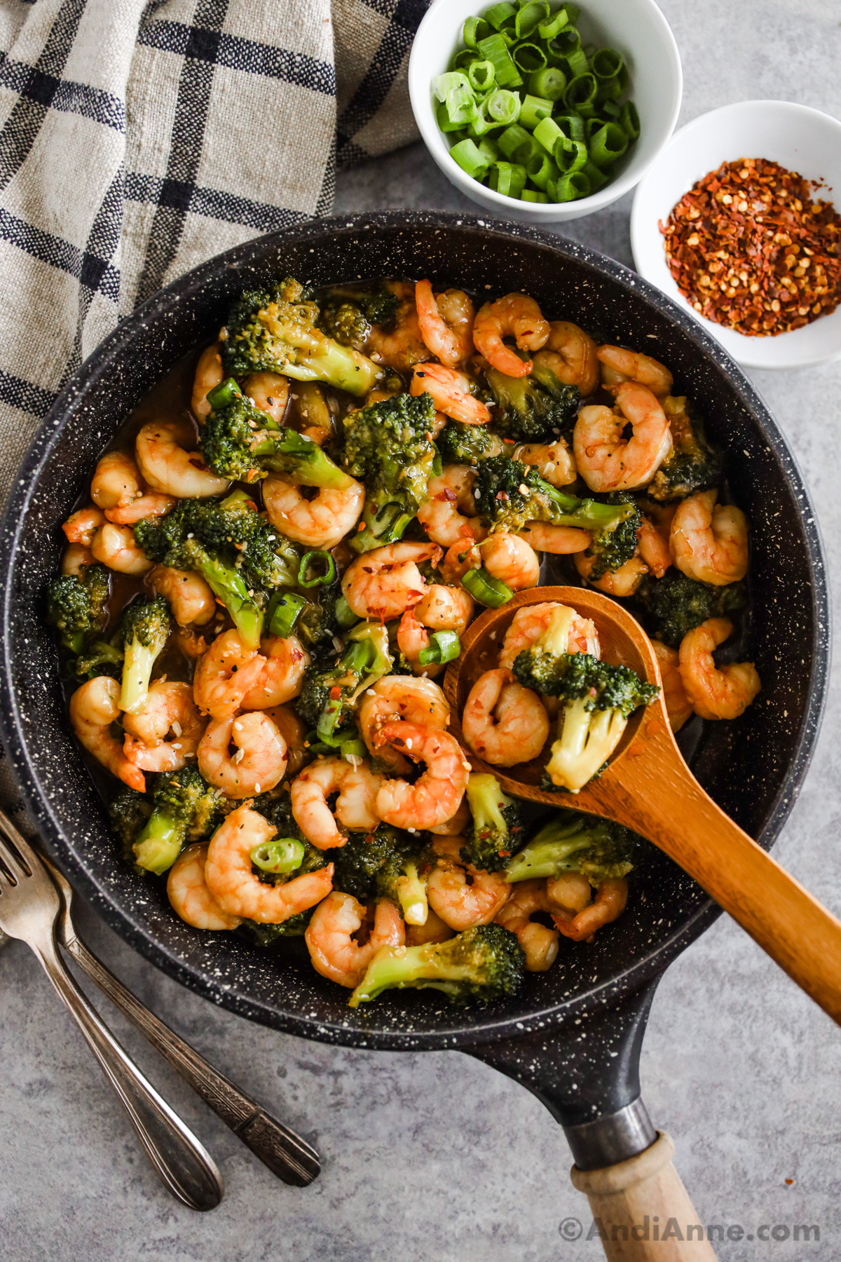 A frying pan with chopped broccoli and shrimp in a honey garlic sauce with bowls of green onion and red pepper flakes.