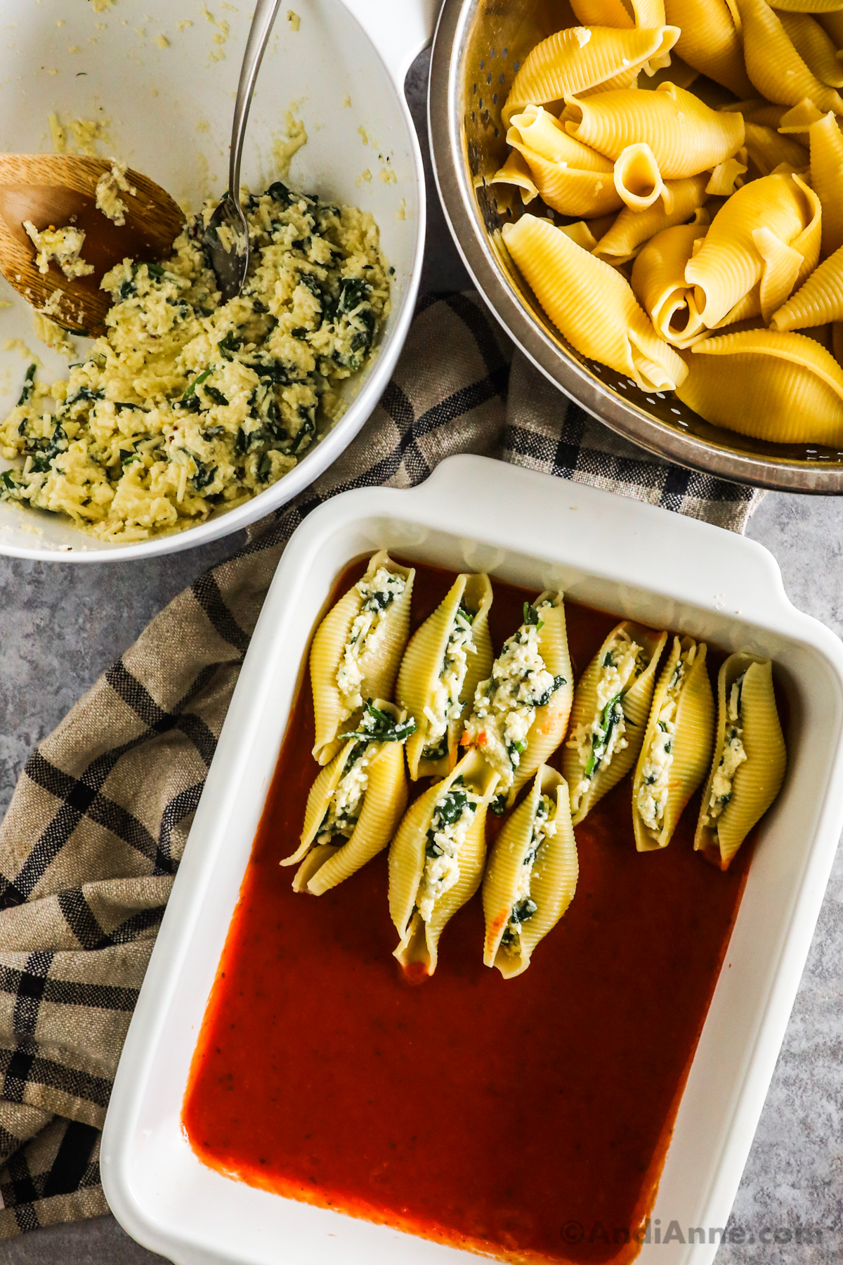 A bowl of spinach ricotta filling, strainer with cooked jumbo shells, and baking dish with marinara sauce and a few stuffed jumbo shells in a single layer.
