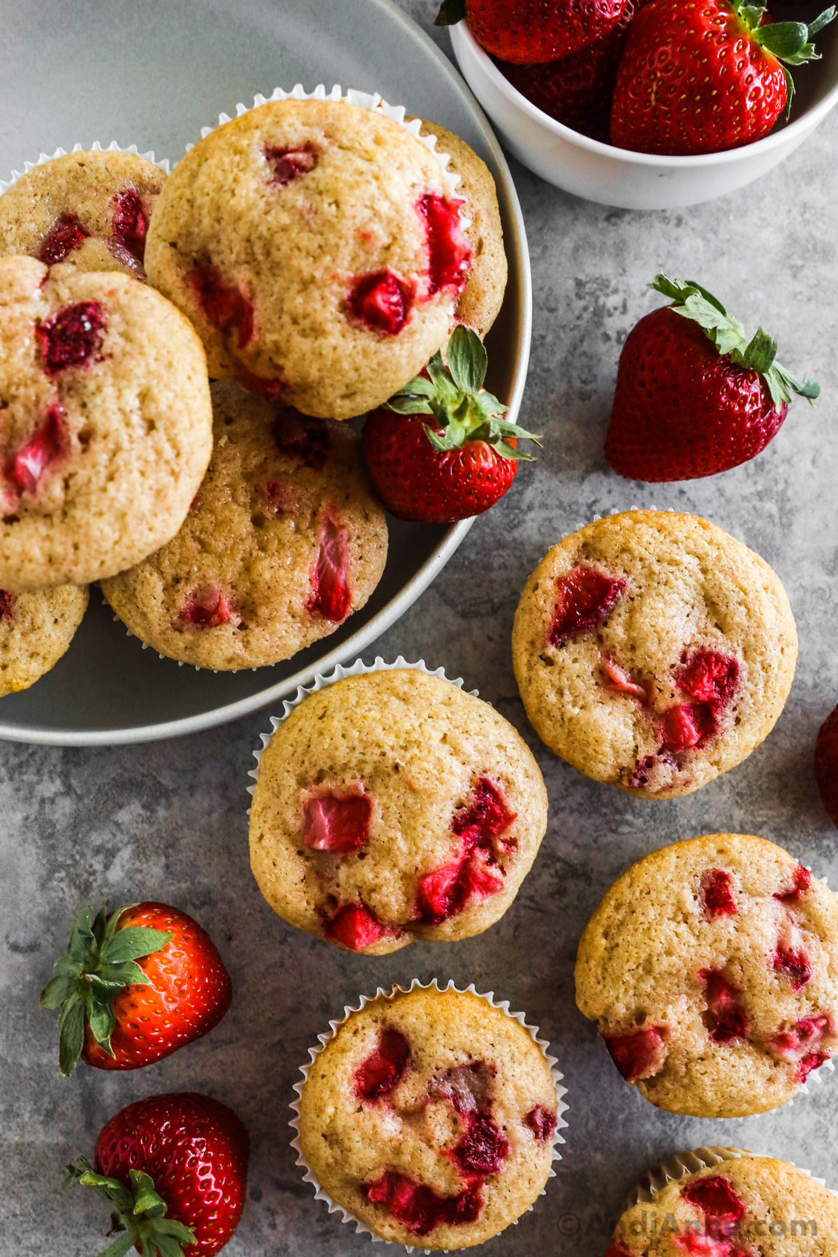 A bunch of strawberry muffins with fresh strawberries surrounding them.