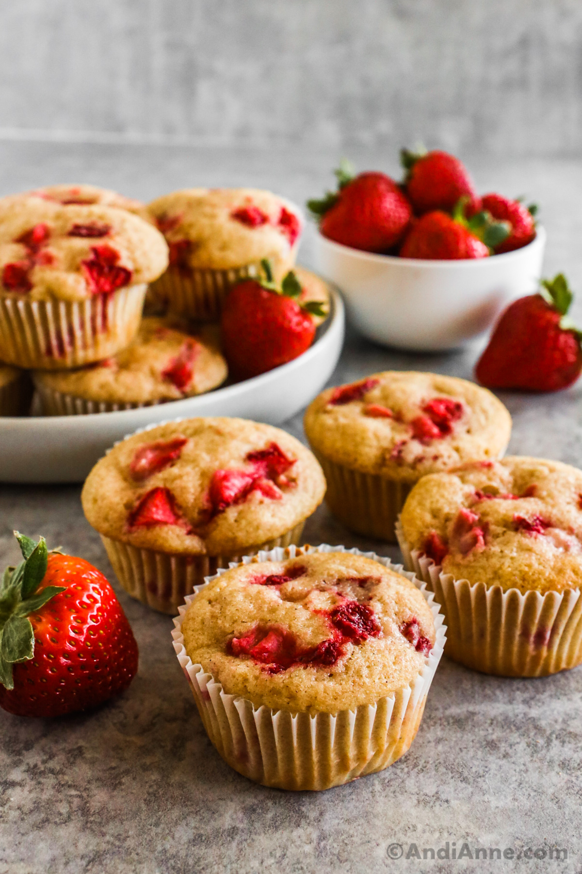 Strawberry muffins on the counter with a bowl of stacked muffins plus fresh strawberries behind them.