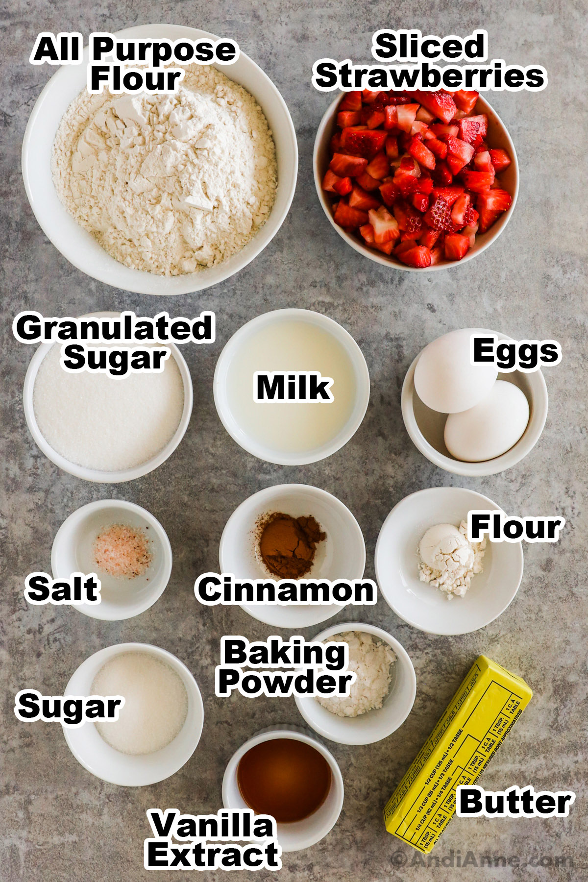 Recipe ingredients on the counter including a bowl of flour, sliced strawberries, sugar, milk, eggs, cinnamon, flour, sugar, baking powder, vanilla and butter.