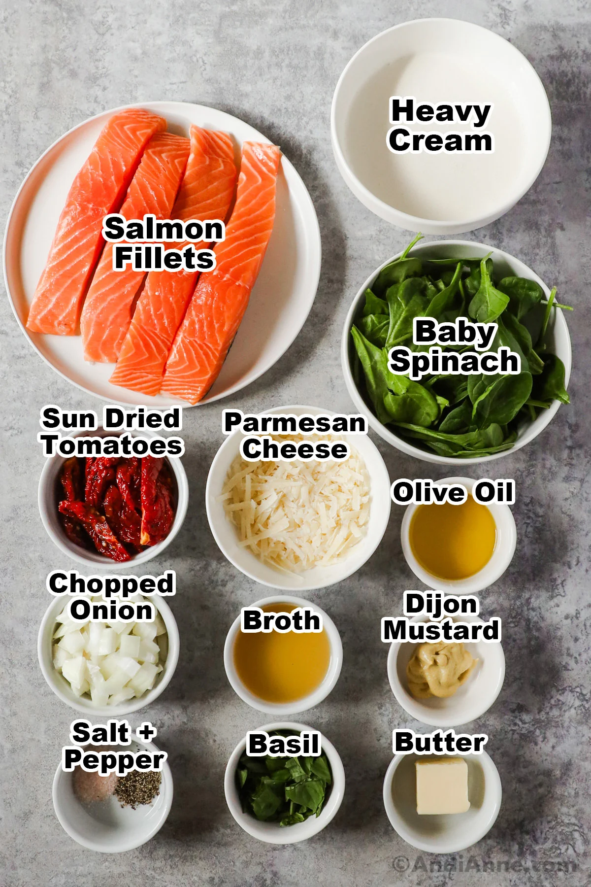 Recipe ingredients in bowls including heavy cream, baby spinach, sun dried tomatoes, parmesan cheese, salmon fillets, olive oil, onion, broth, spices and basil.