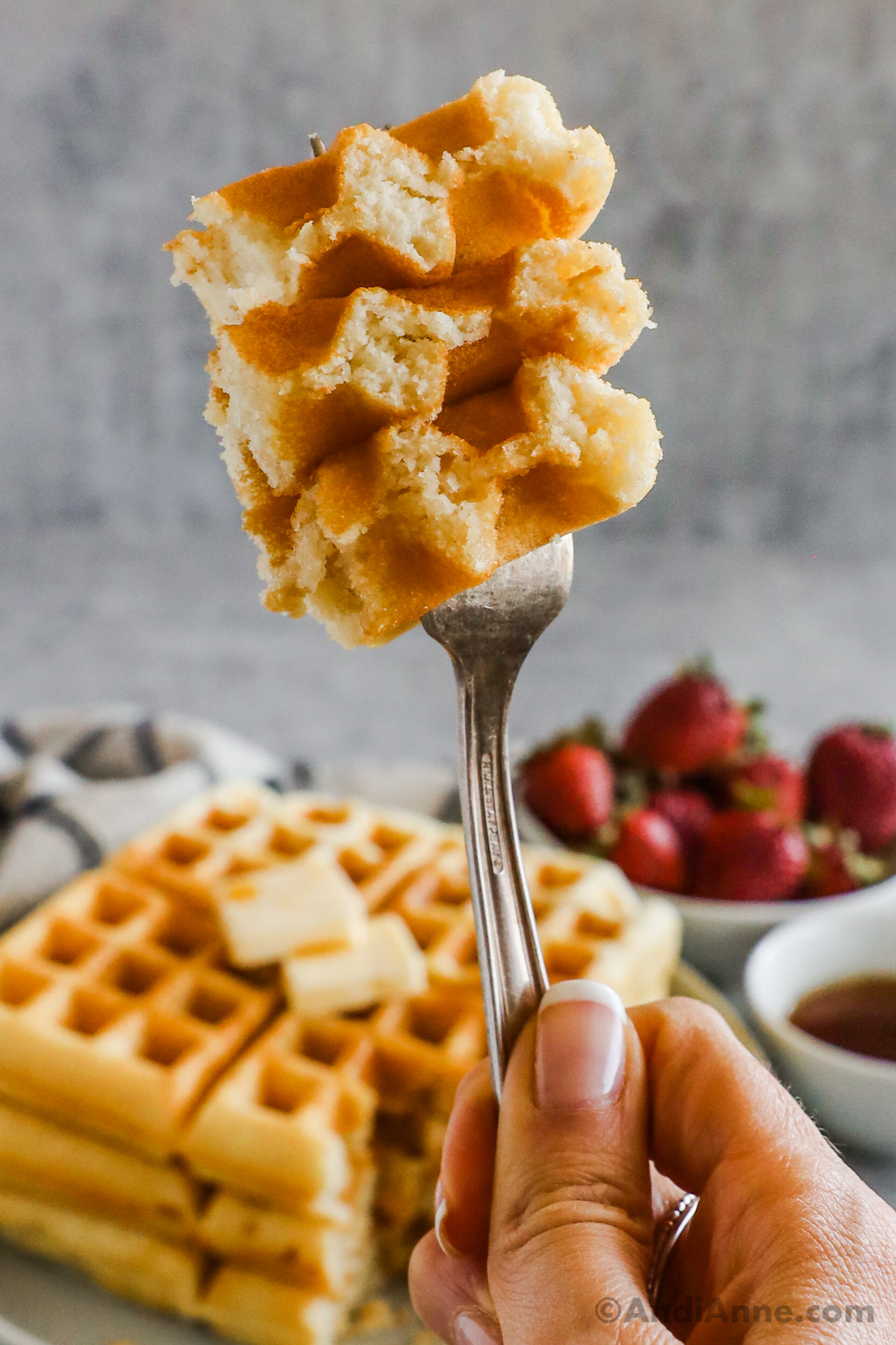 A hand holding a fork with a bite of waffles. Stack of waffles and bowl of strawberries in the background.