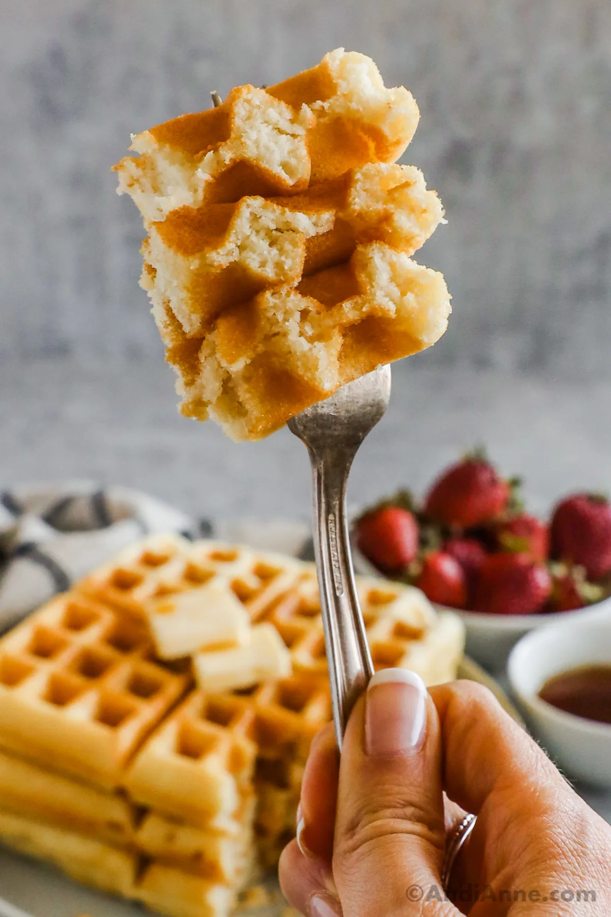 A hand holding a fork with a bite of waffles. Stack of waffles and bowl of strawberries in the background.