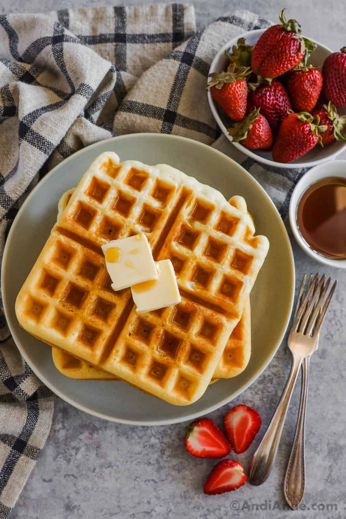 Classic waffle recipe on a plate with butter and drizzled with syrup, surrounded by forks, a bowl of strawberries and a bowl of syrup.