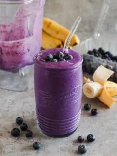 Glass of blueberry smoothie with banana and blender in background.