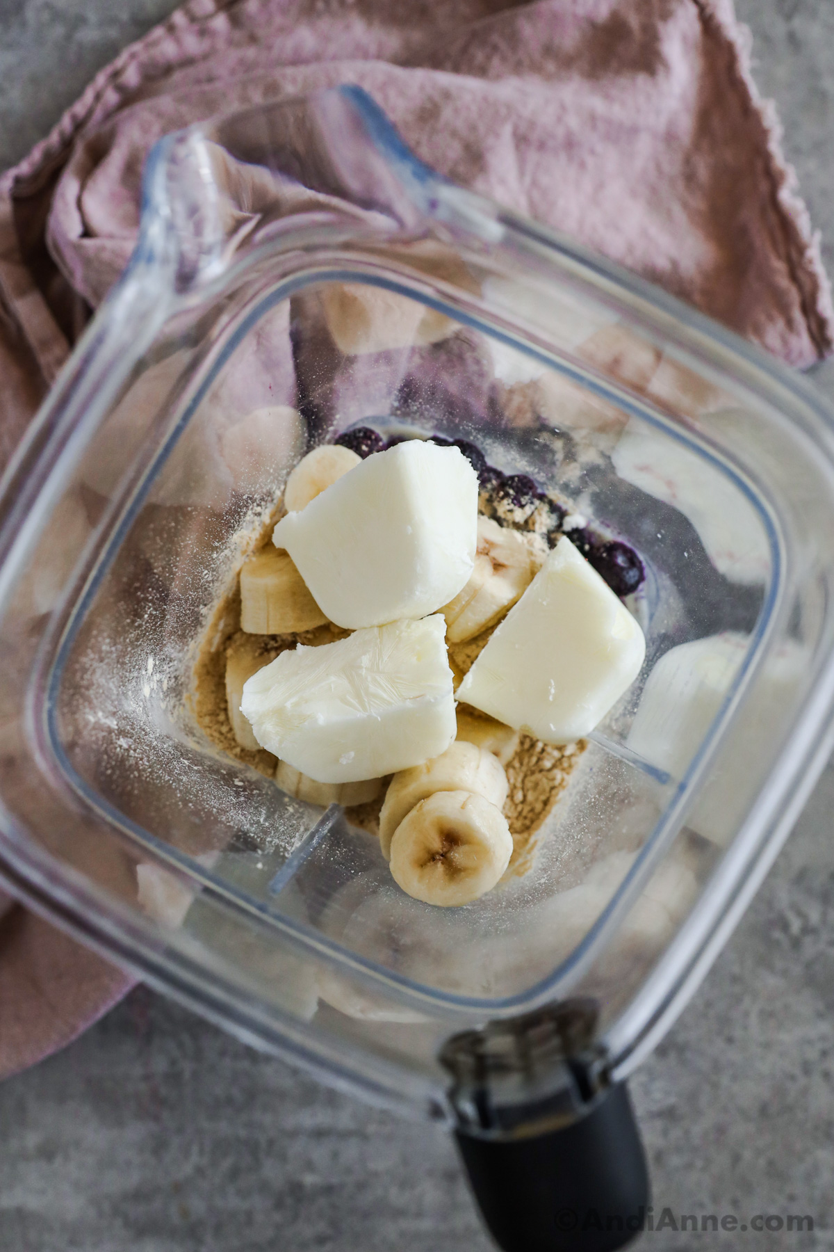 Looking into a blender cup with cubes of frozen yogurt, sliced banana, blueberries and protein powder.