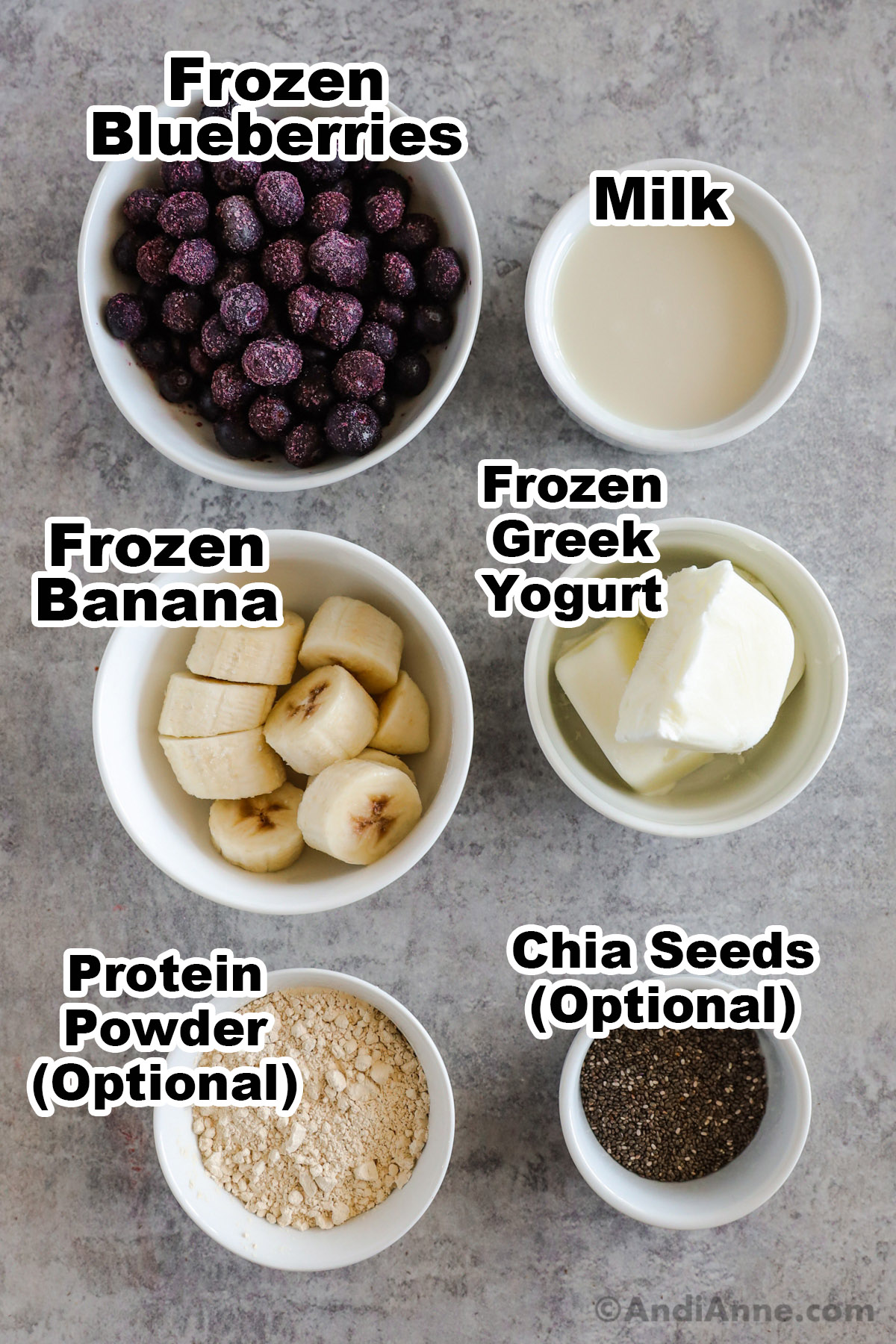 Recipe ingredients in bowls, including frozen banana, milk, frozen blueberries and cubes of frozen greek yogurt, proien powder and chia seeds.