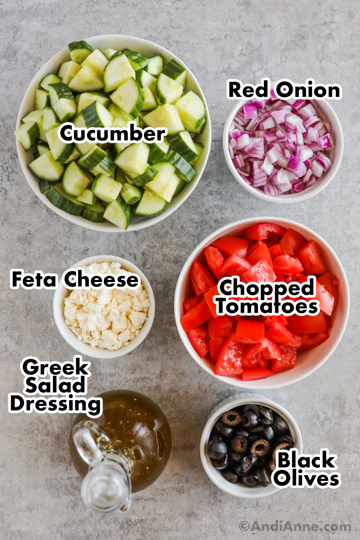 Bowls of chopped cucumber, onion, tomato, feta cheese, sliced olives, and jar of greek salad dressing.