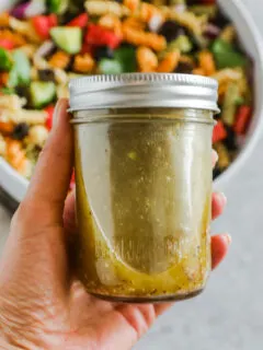 Hand holding a mason jar with greek salad dressing with bowl of pasta salad in background.