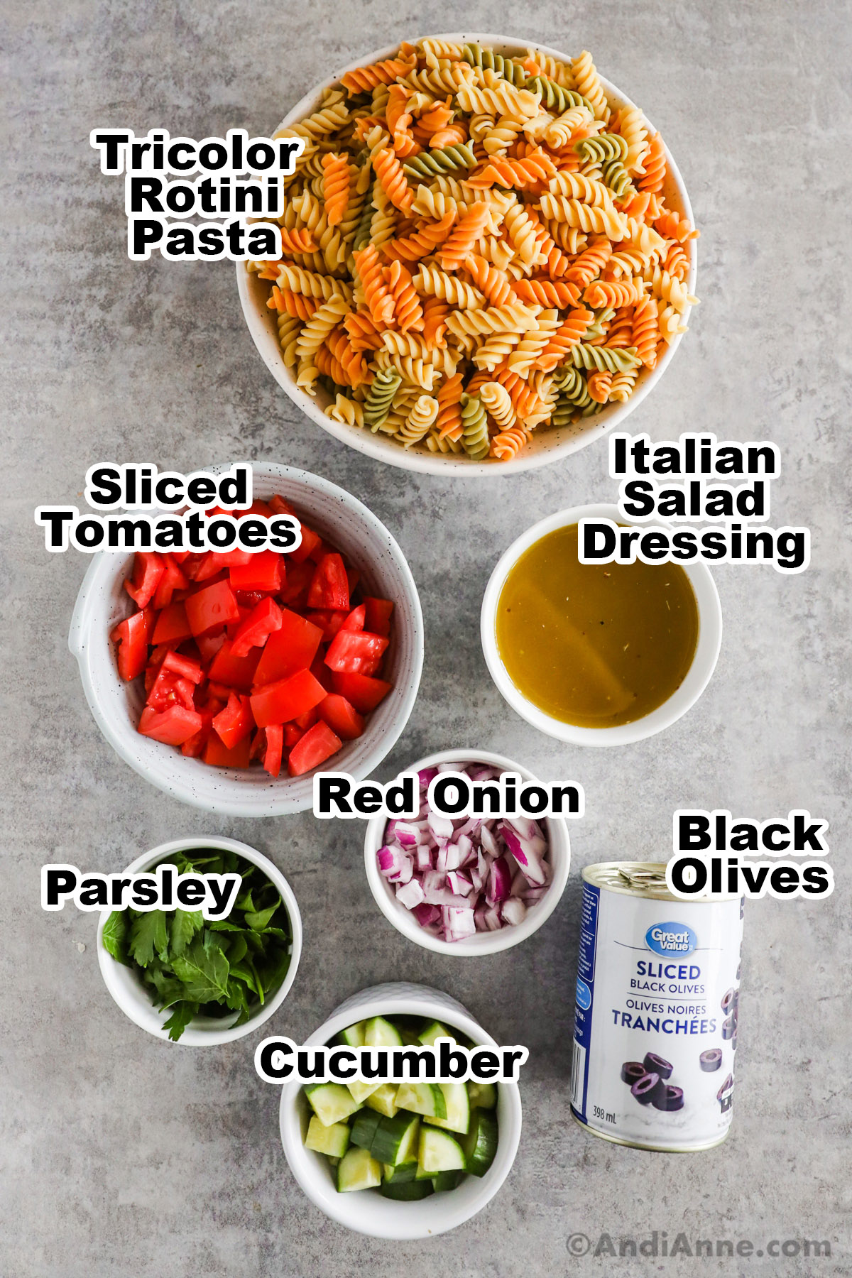 Recipe ingredients on a counter including a bowl of rotini pasta, bowls of tomatoes, salad dressing, red onion, parsley, cucumber and canned black olives.