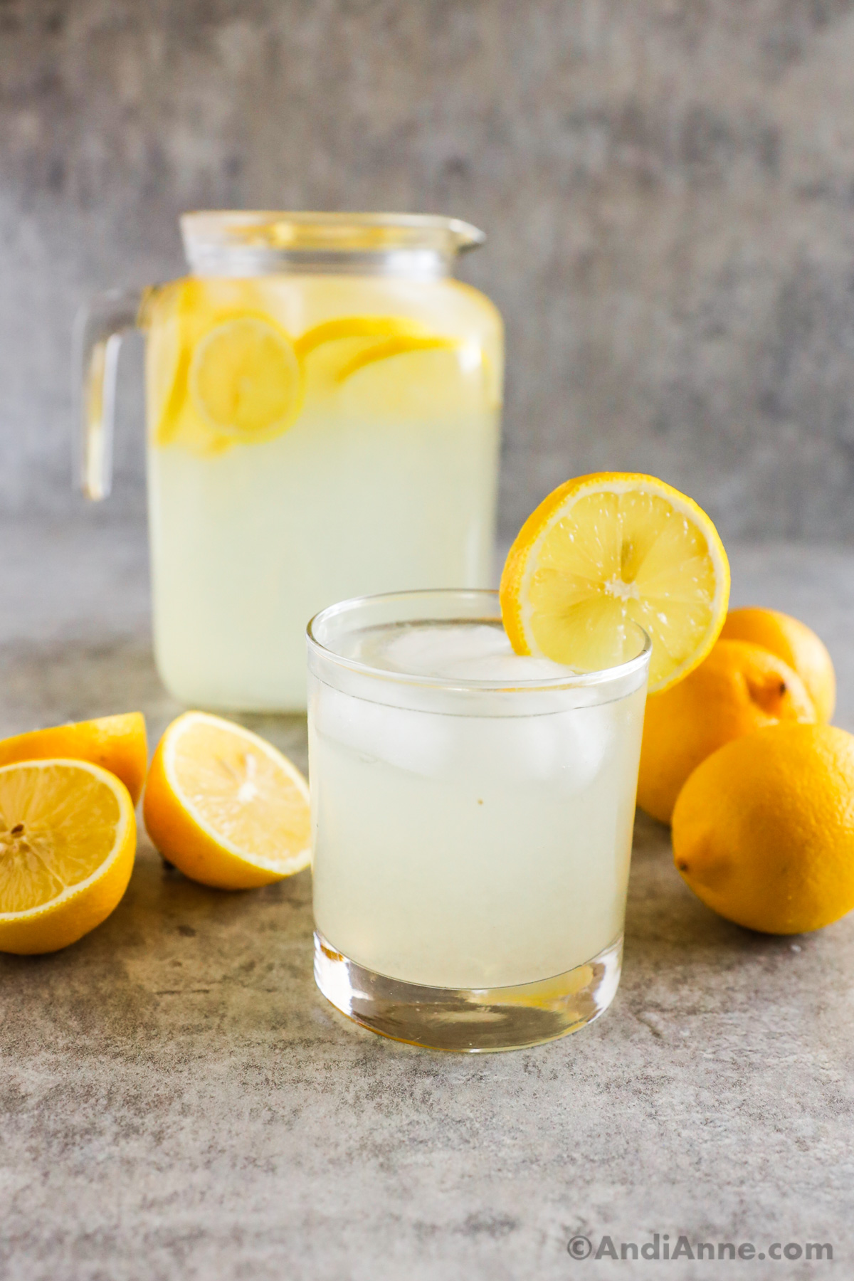 a full glass of lemonade with ice and a slice of lemon as garnish on edge of glass. Lemons and full pitcher of lemonade sit in the background