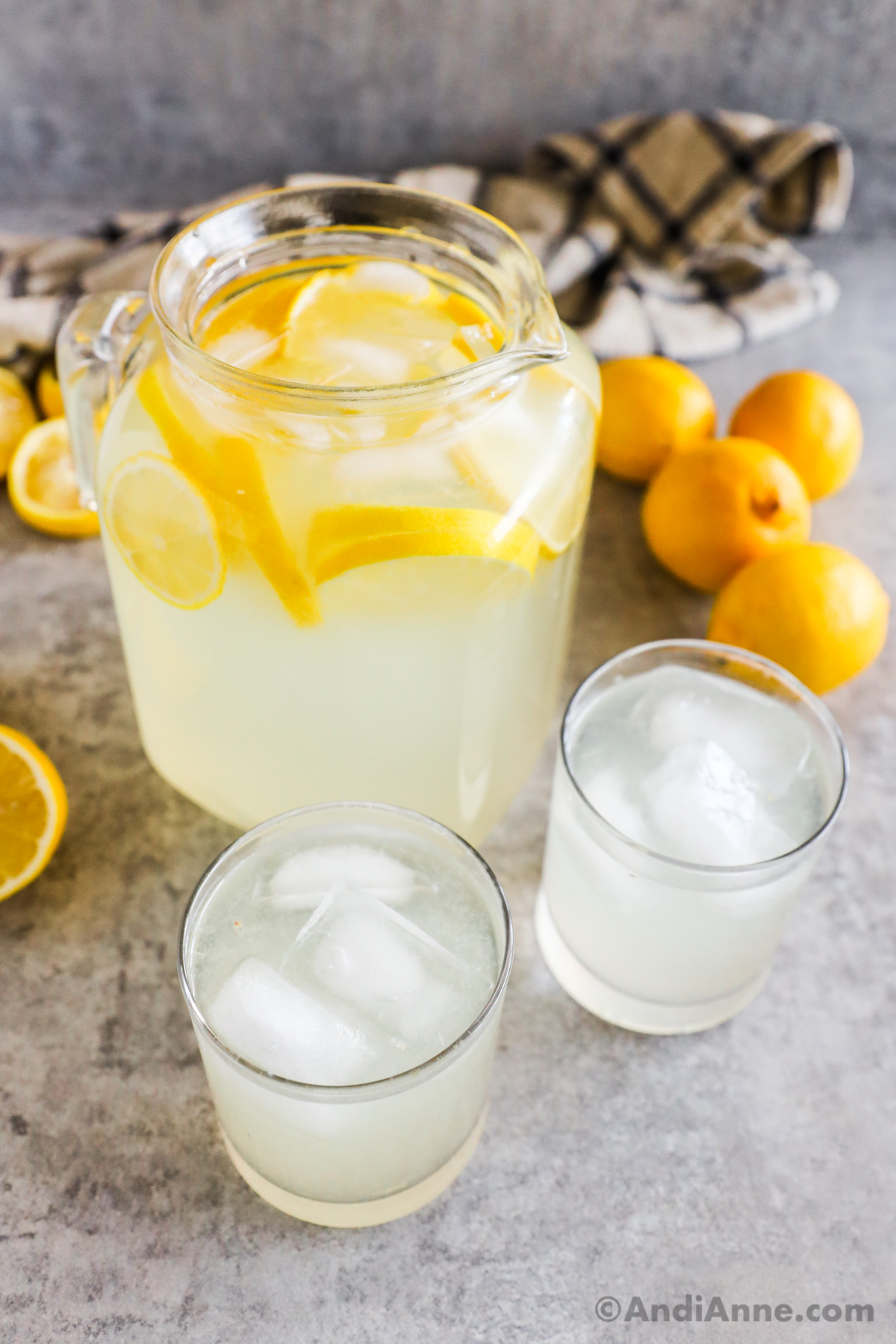 A 1.5 litre glass pitcher full of lemonade with lemon slices behind two glasses of lemonade with ice cubes. Whole and sliced lemons in background.