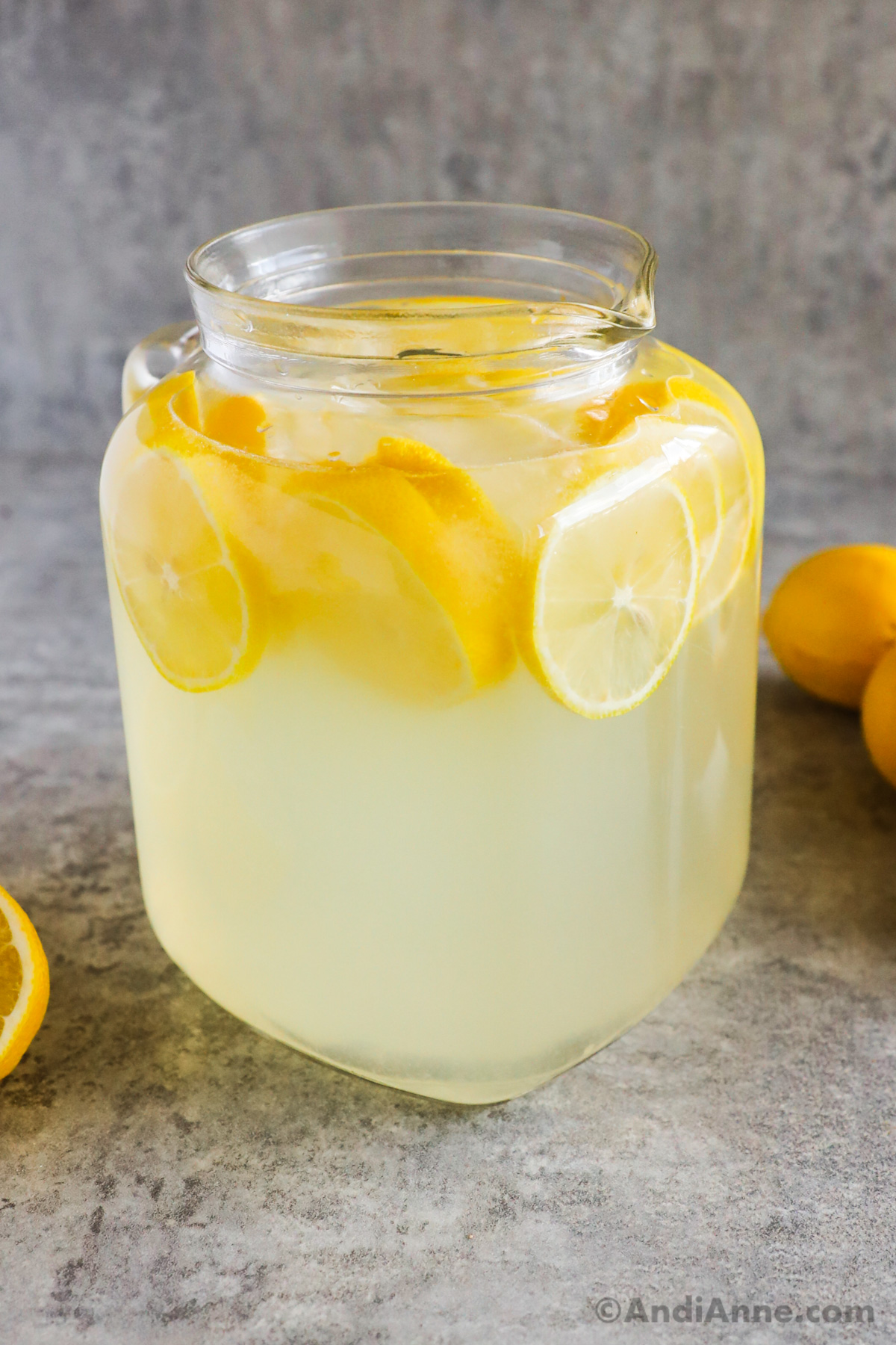 closeup of a full glass pitcher of lemonade with sliced lemons inside. whole and sliced lemons sit in background