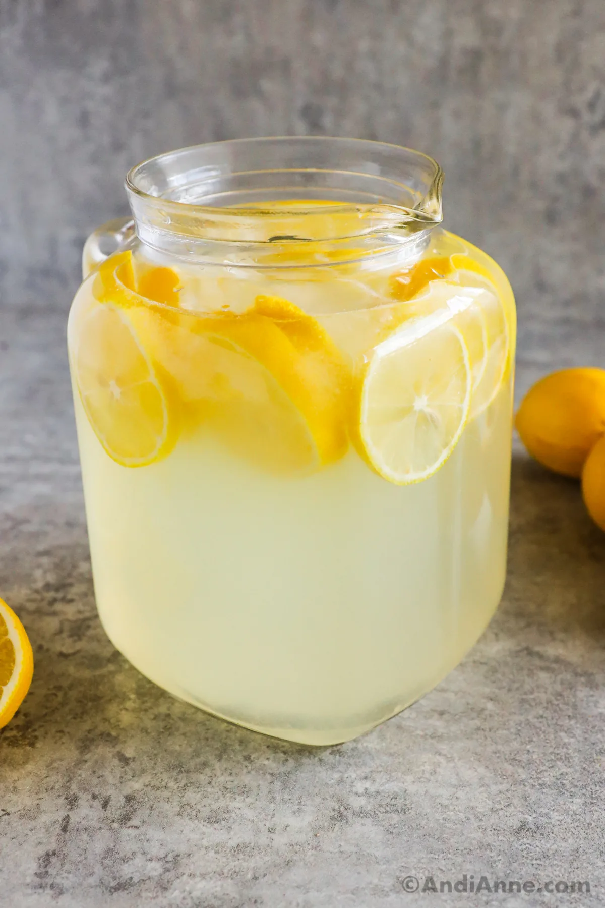 closeup of a full glass pitcher of lemonade with sliced lemons inside. whole and sliced lemons sit in background