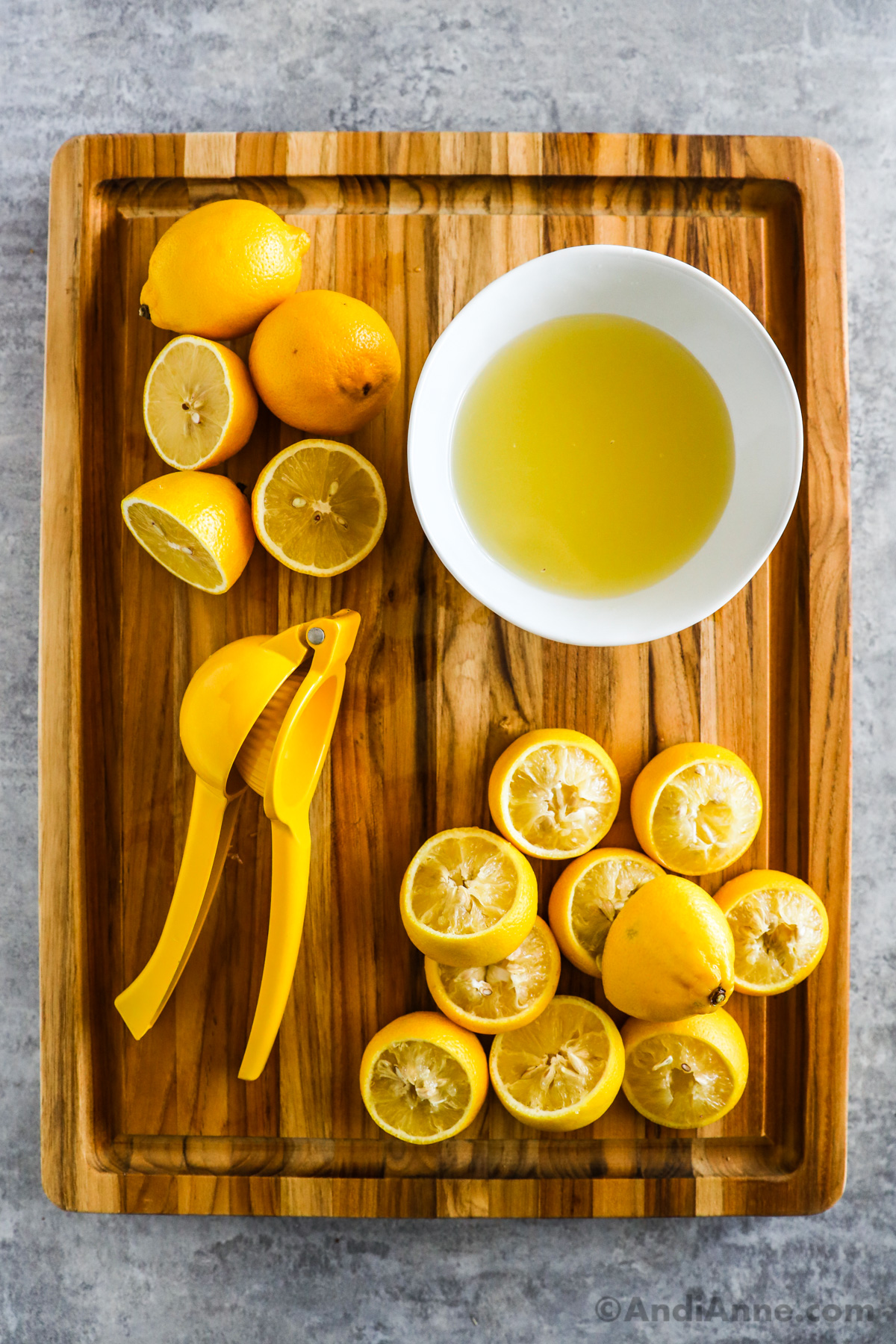 A bowl of lemon juice, sliced lemons and a yellow hand-held juicer sit on a wooden cutting board.