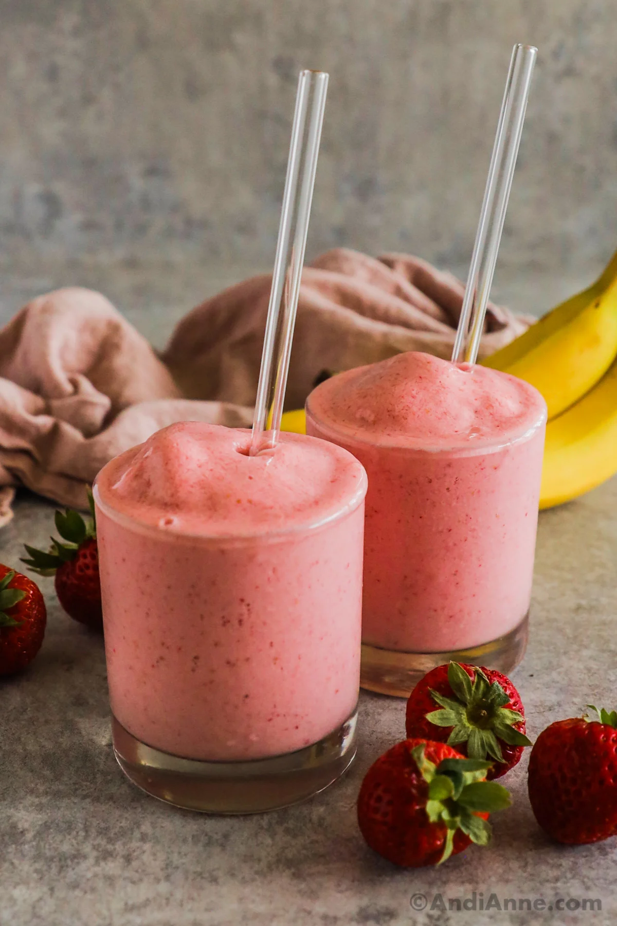Two glasses of strawberry banana smoothie with straws surrounded by bananas and fresh strawberries.
