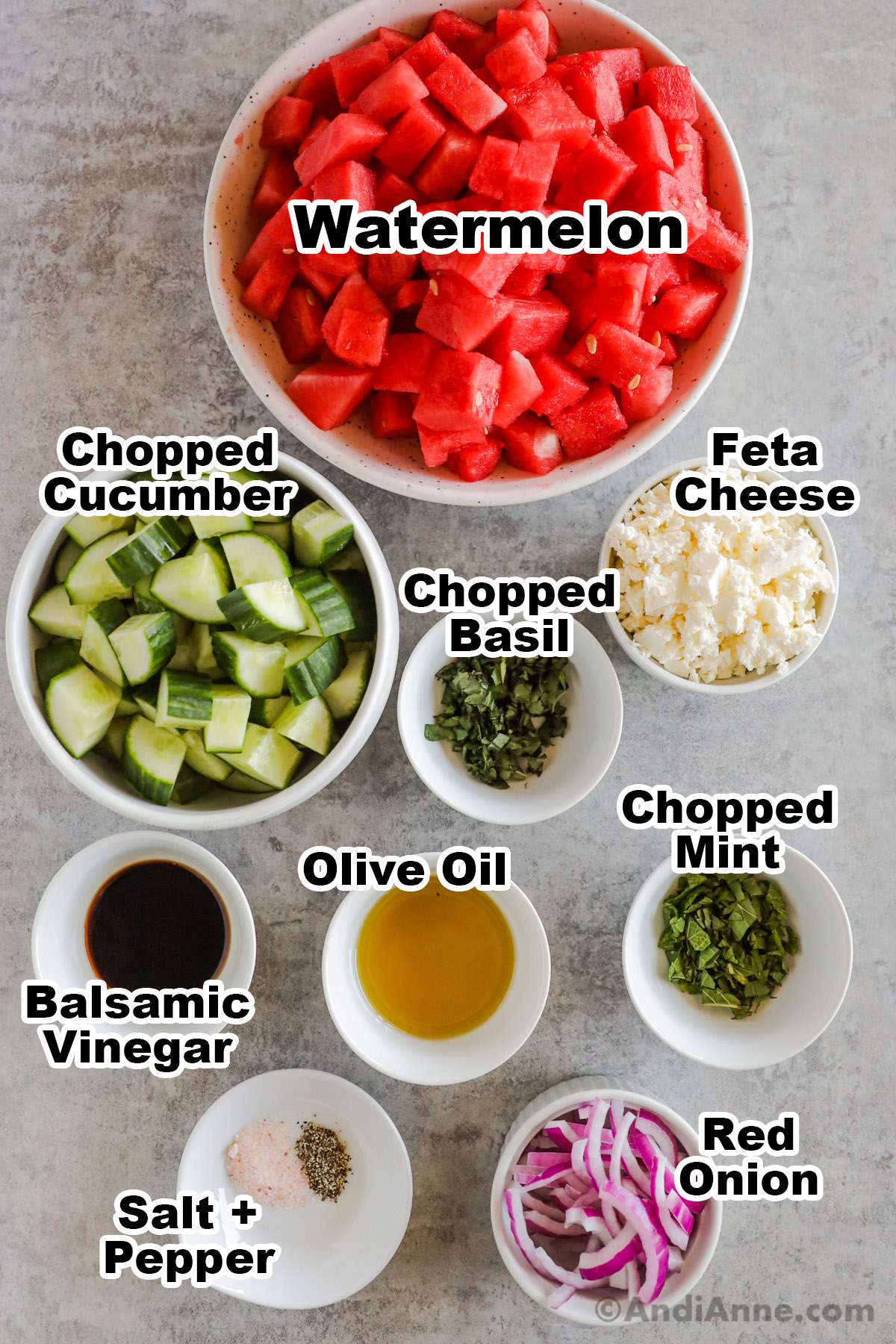 Recipe ingredients in bowls including cubed watermelon, chopped cucumber, feta cheese, chopped basil and mint, balsamic vinegar, olive oil, and red onion.
